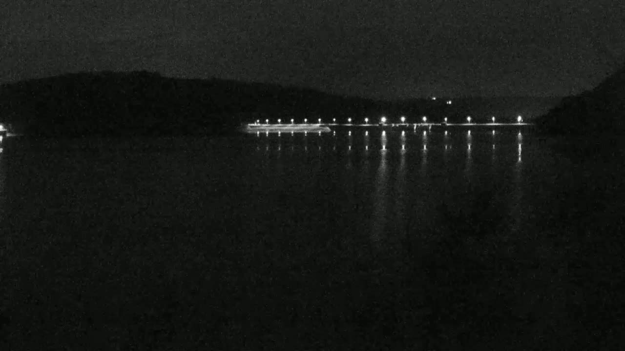 A nighttime view of a calm body of water reflecting the lights of a structure along the far shore, with dark hills visible in the background.