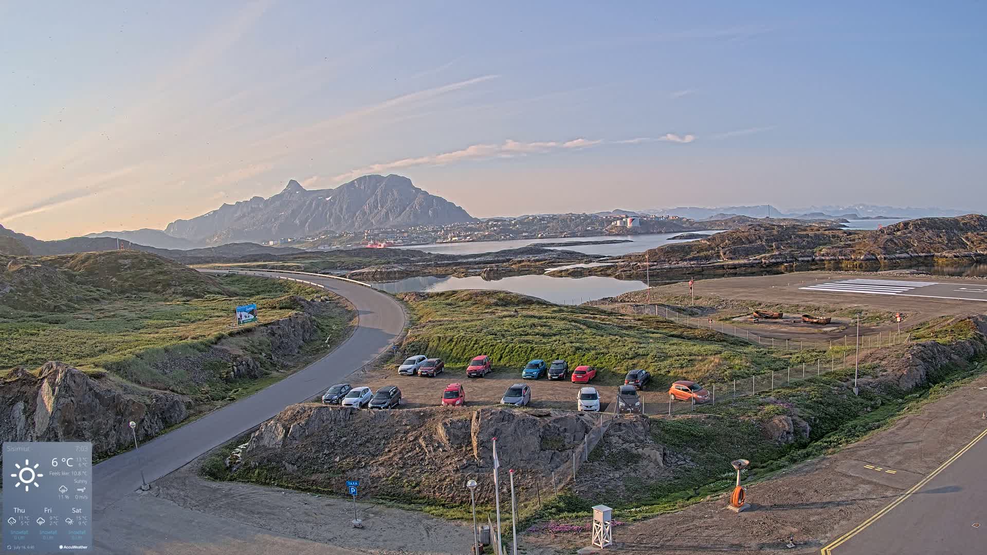 A small airport with a parking lot full of cars overlooks a calm body of water and mountainous landscape under sunny skies.