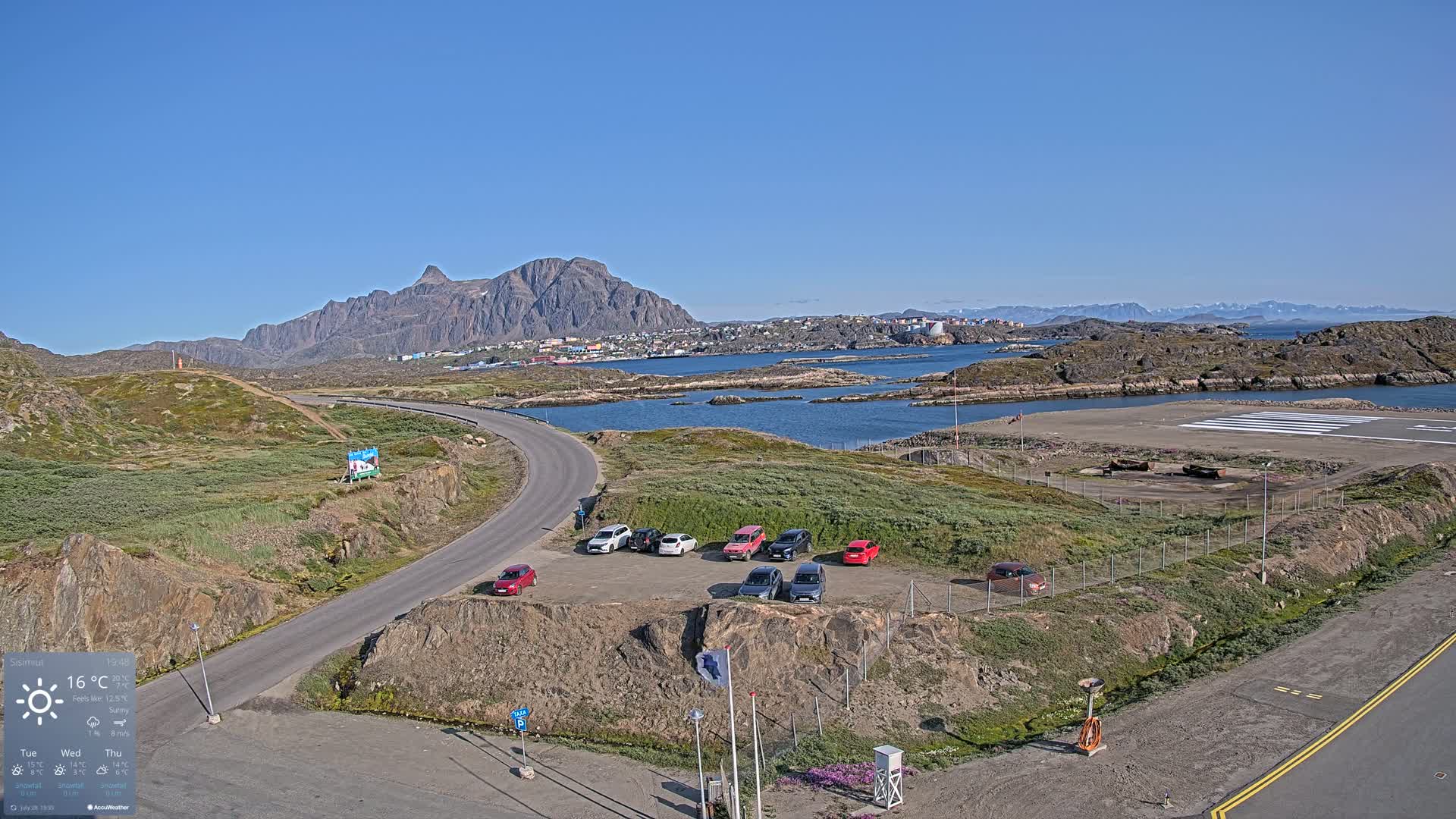 A sunny day reveals a small town nestled in a rocky bay near a small airport, with several cars parked nearby and mountains in the background.