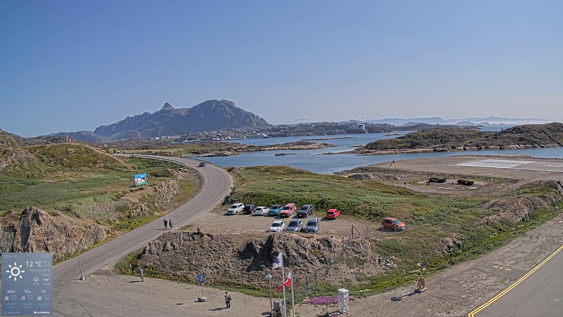 A sunny day reveals a small town nestled in a rocky bay near a small airport, with several cars parked nearby and mountains in the background.