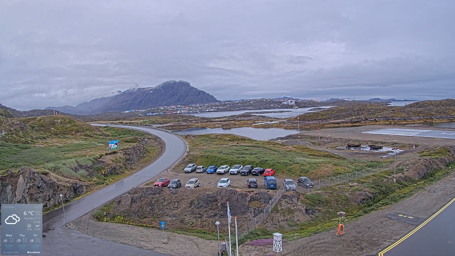 A small airport with a parking lot full of cars is situated near a town on a cloudy day, with a mountain in the background.