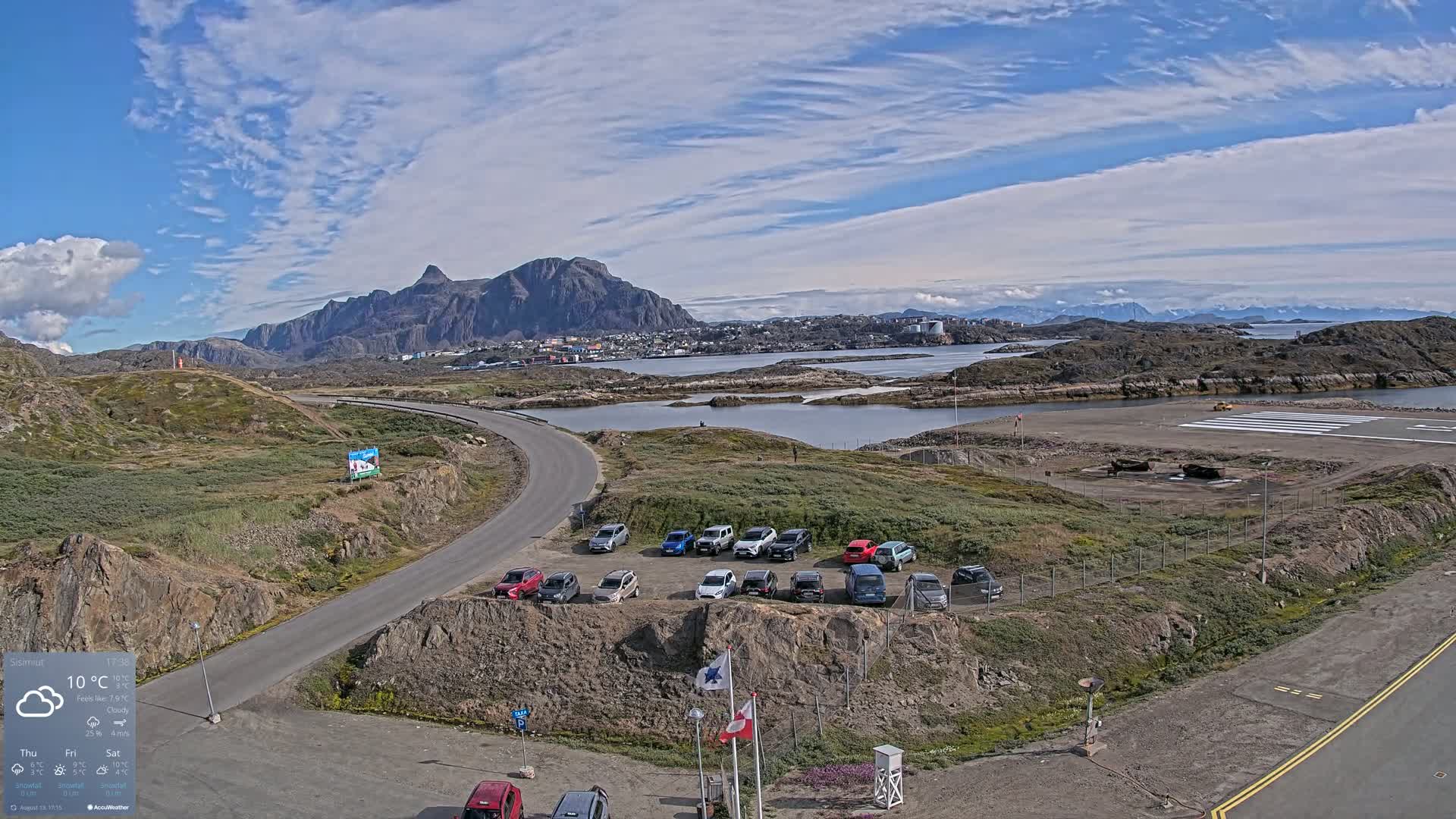 A partly cloudy sky overlooks a small town nestled between mountains and a body of water, with a small airport and a parking lot of cars nearby.