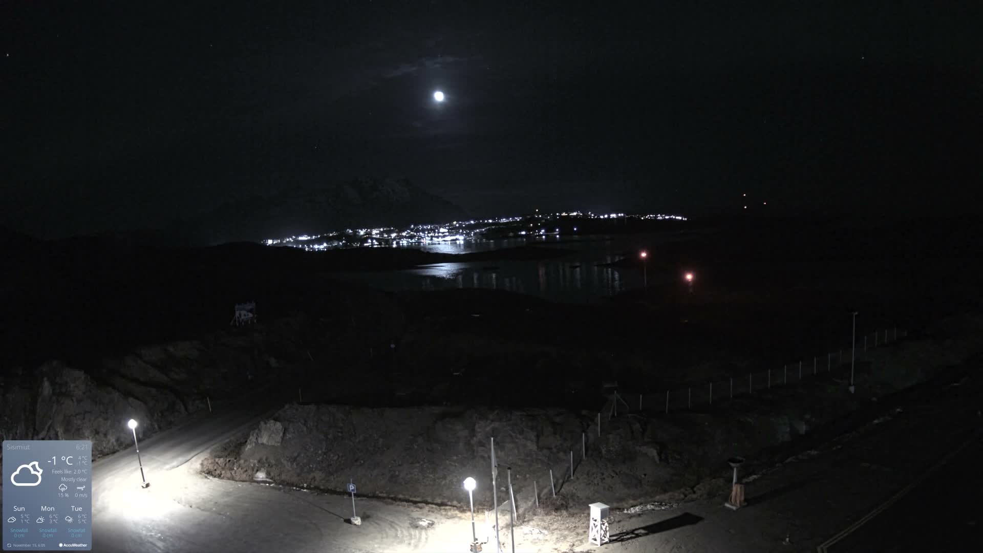 A dark, moonlit night scene showcases a coastal town with twinkling lights reflected on a body of water, flanked by dark mountains and a foreground of winding roads illuminated by streetlights, under a mostly clear sky.