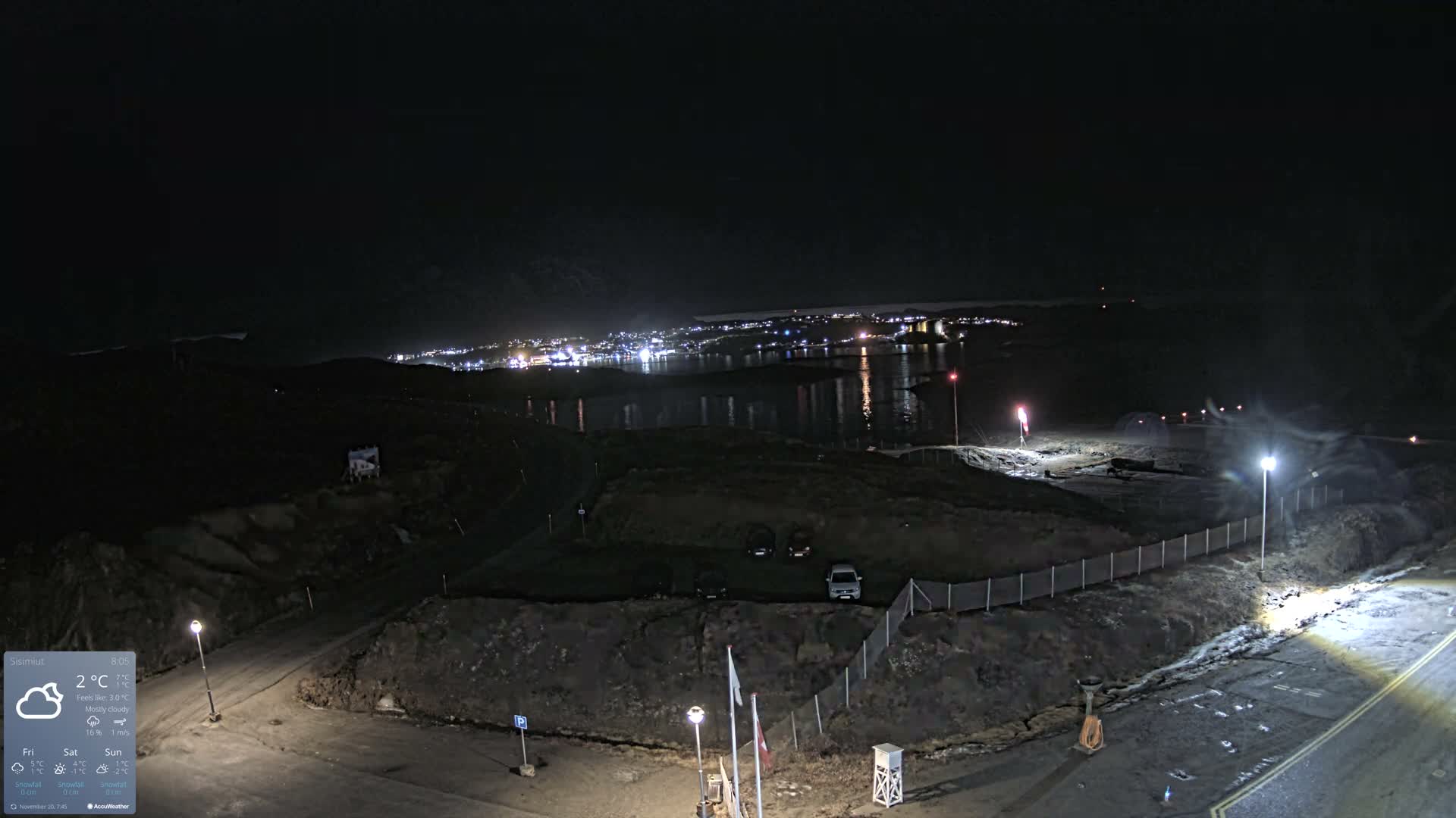 A wide-angle nighttime view shows a rugged foreground with illuminated roads, streetlights, and a red windsock, overlooking a calm body of water reflecting the bright lights of a distant town or city under a dark night sky.
