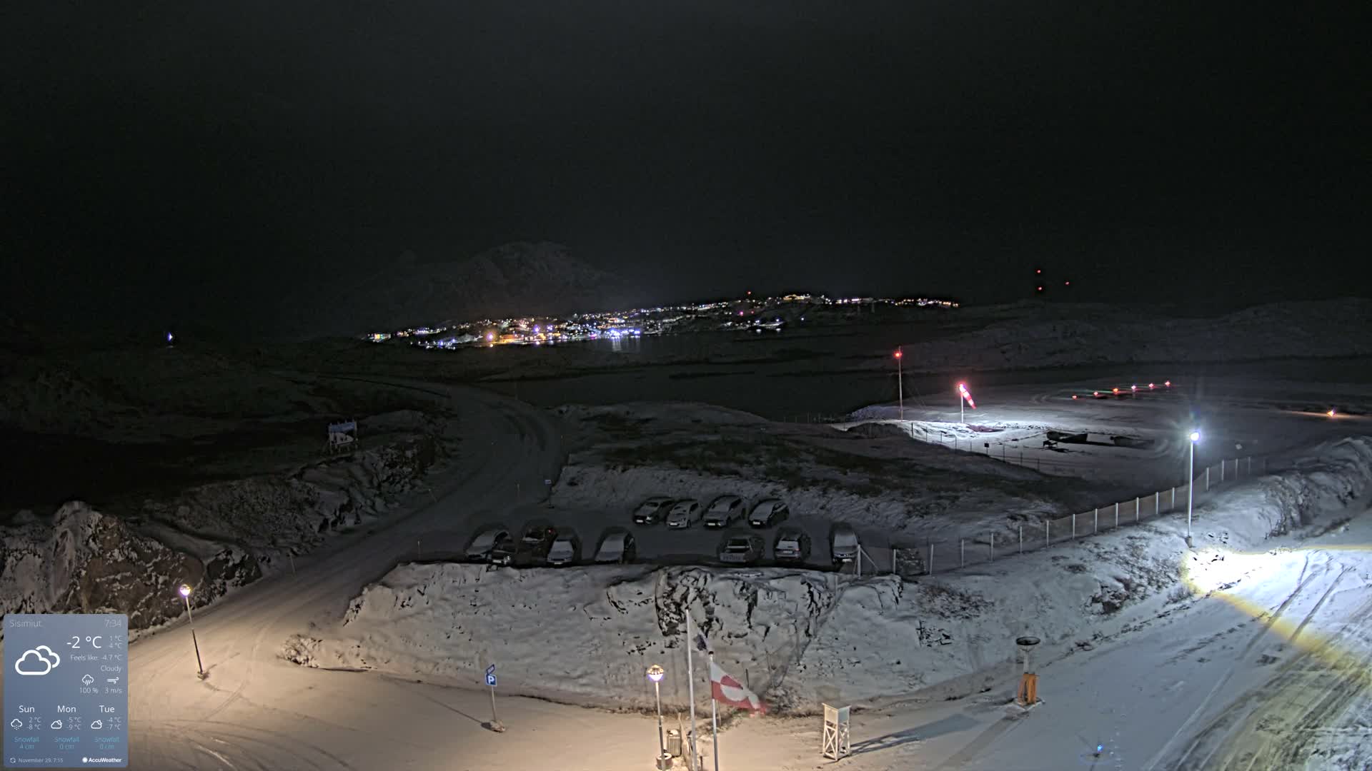A panoramic night view captures a snow-covered, hilly landscape featuring a brightly lit town across a dark bay and an illuminated airport runway, all under a cloudy winter sky.