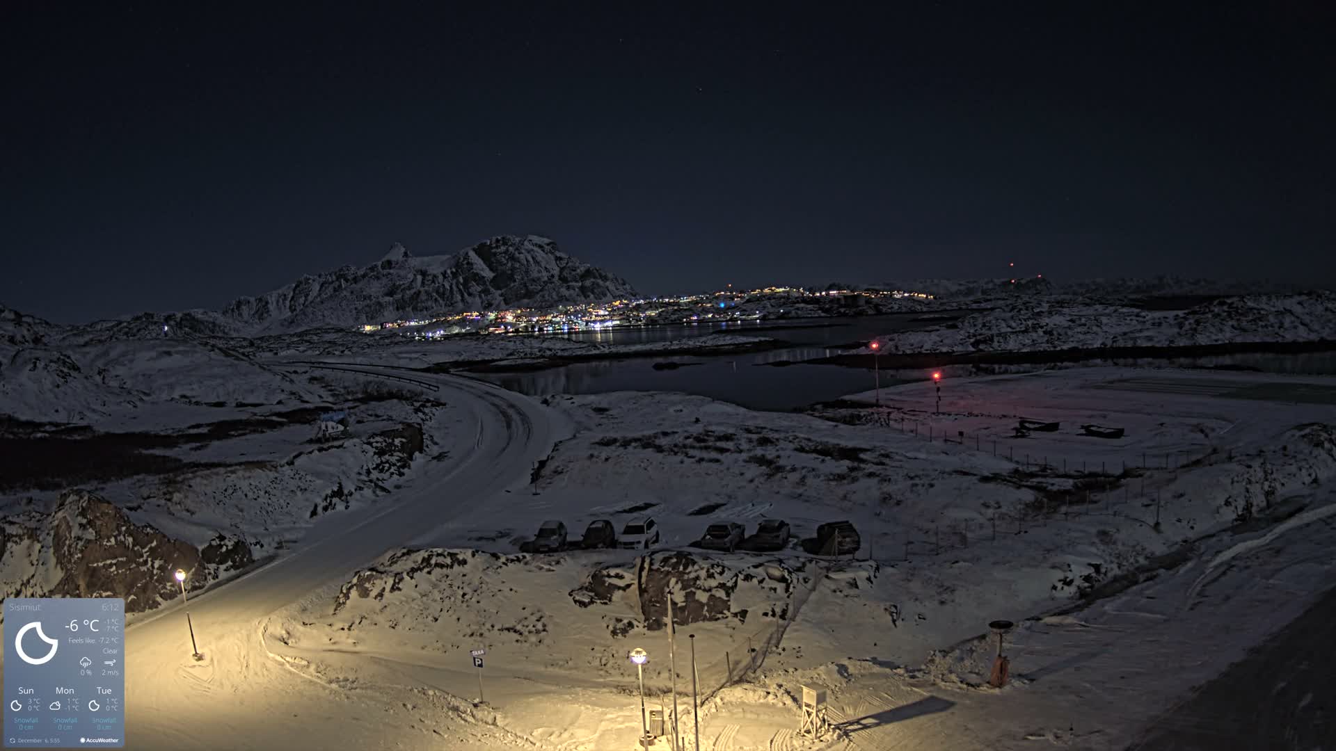 A vast, snow-covered mountainous landscape is visible at night under a clear, star-filled sky, with a brightly lit town nestled along a body of water, a winding road, and a parking area illuminated in the foreground.
