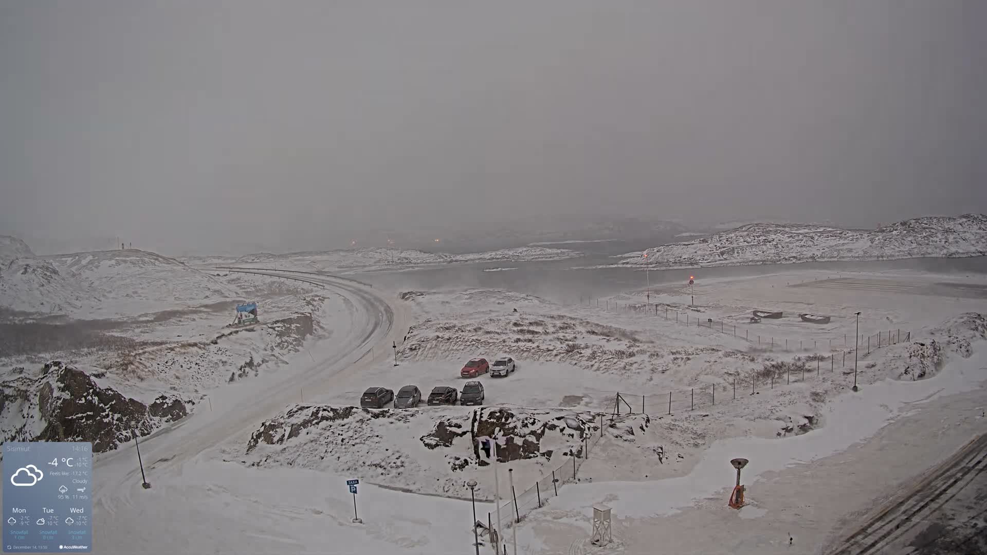 A vast, snow-covered mountainous landscape is visible at night under a clear, star-filled sky, with a brightly lit town nestled along a body of water, a winding road, and a parking area illuminated in the foreground.