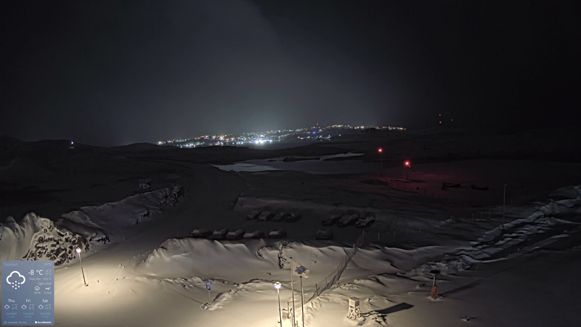 A vast, snow-covered landscape at night is illuminated by a few streetlights in the foreground and the distant glow of a town under a dark, overcast sky.