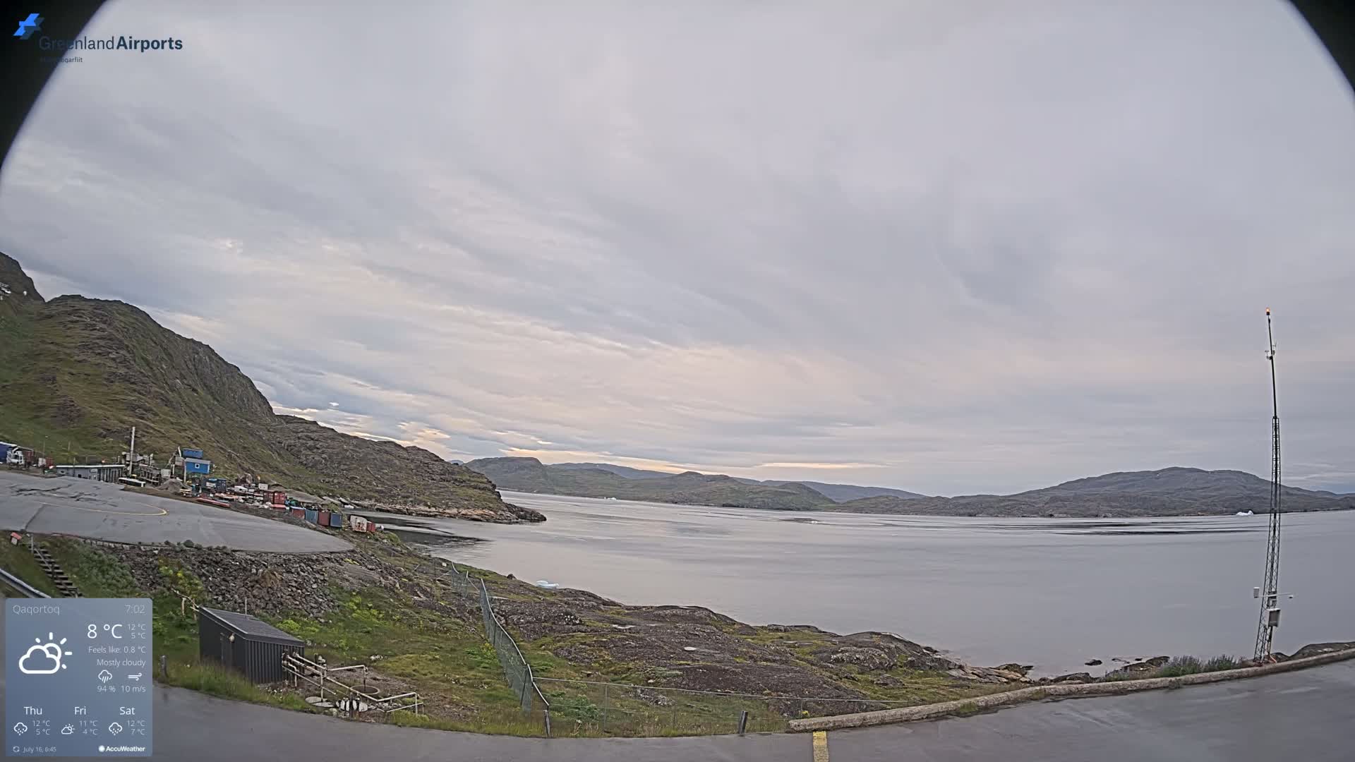 A mostly cloudy day reveals a rocky, coastal landscape with calm water, a small airfield, and a tall communication tower.