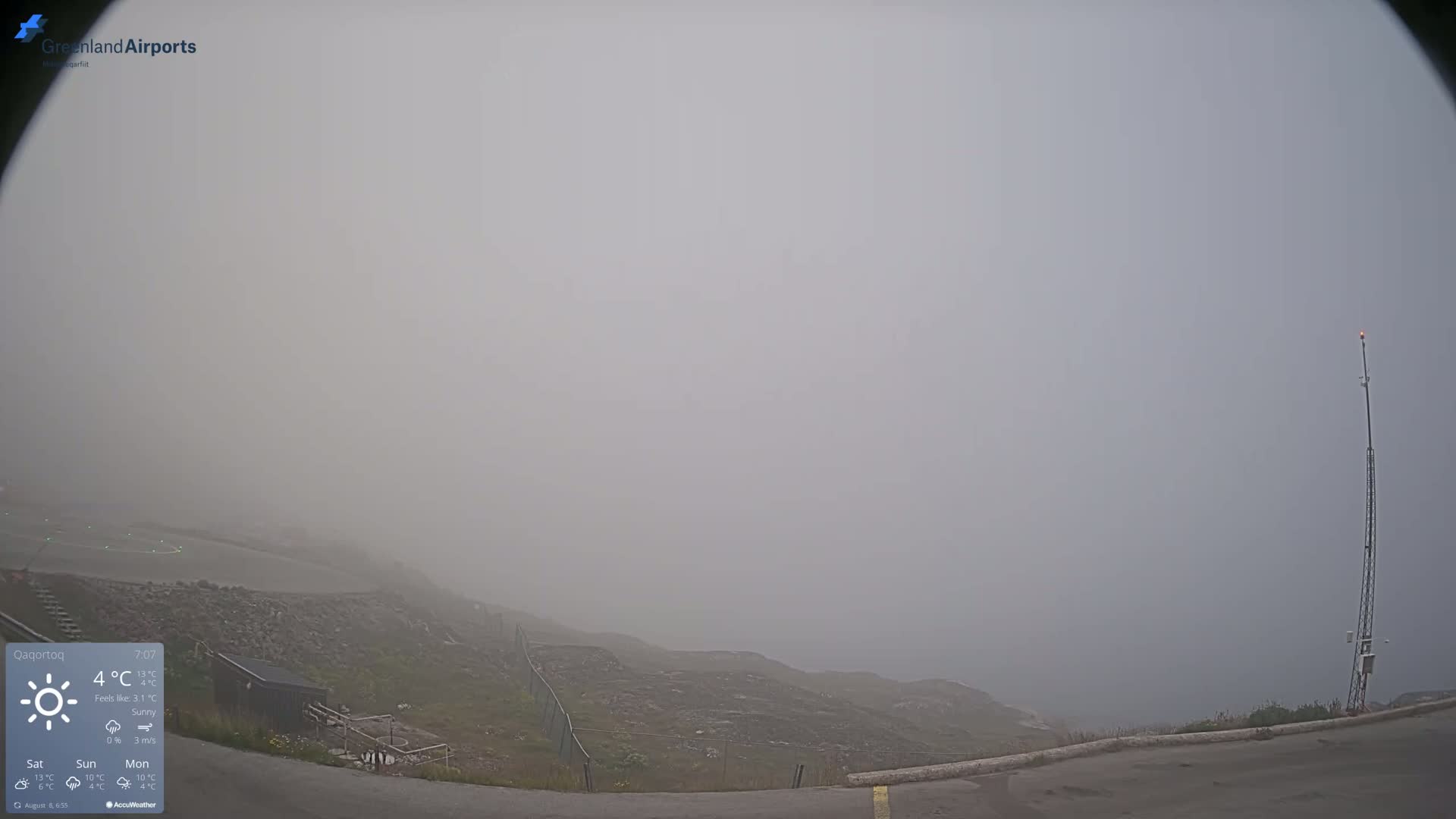 A foggy, outdoor scene shows a rocky, sparsely vegetated hillside with a tall thin tower near a paved road.