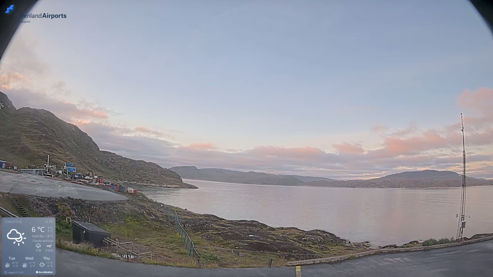 A calm body of water is seen from a rocky shoreline under a mostly clear sky with some pink-hued clouds at dawn.