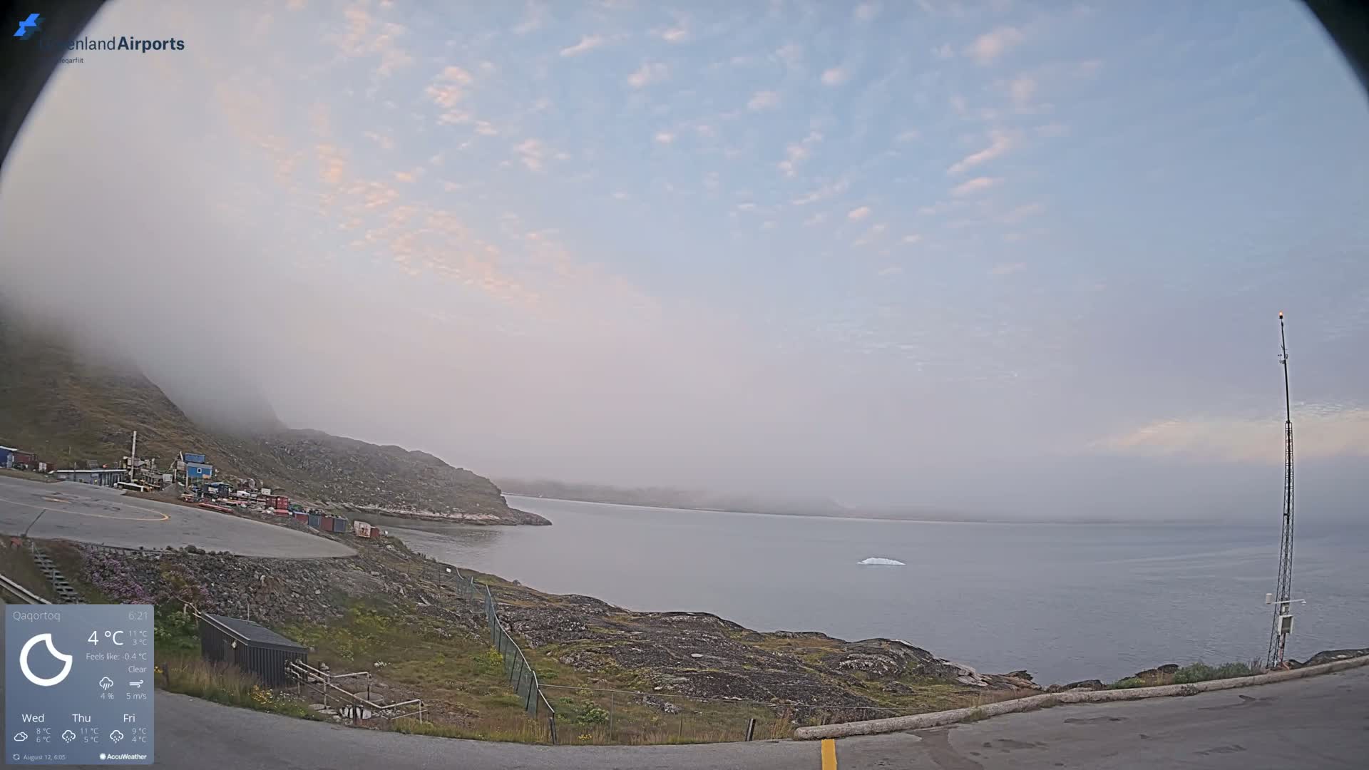 A foggy coastal scene shows a body of water with a single iceberg, a rocky shoreline with sparse vegetation, and a small airfield.
