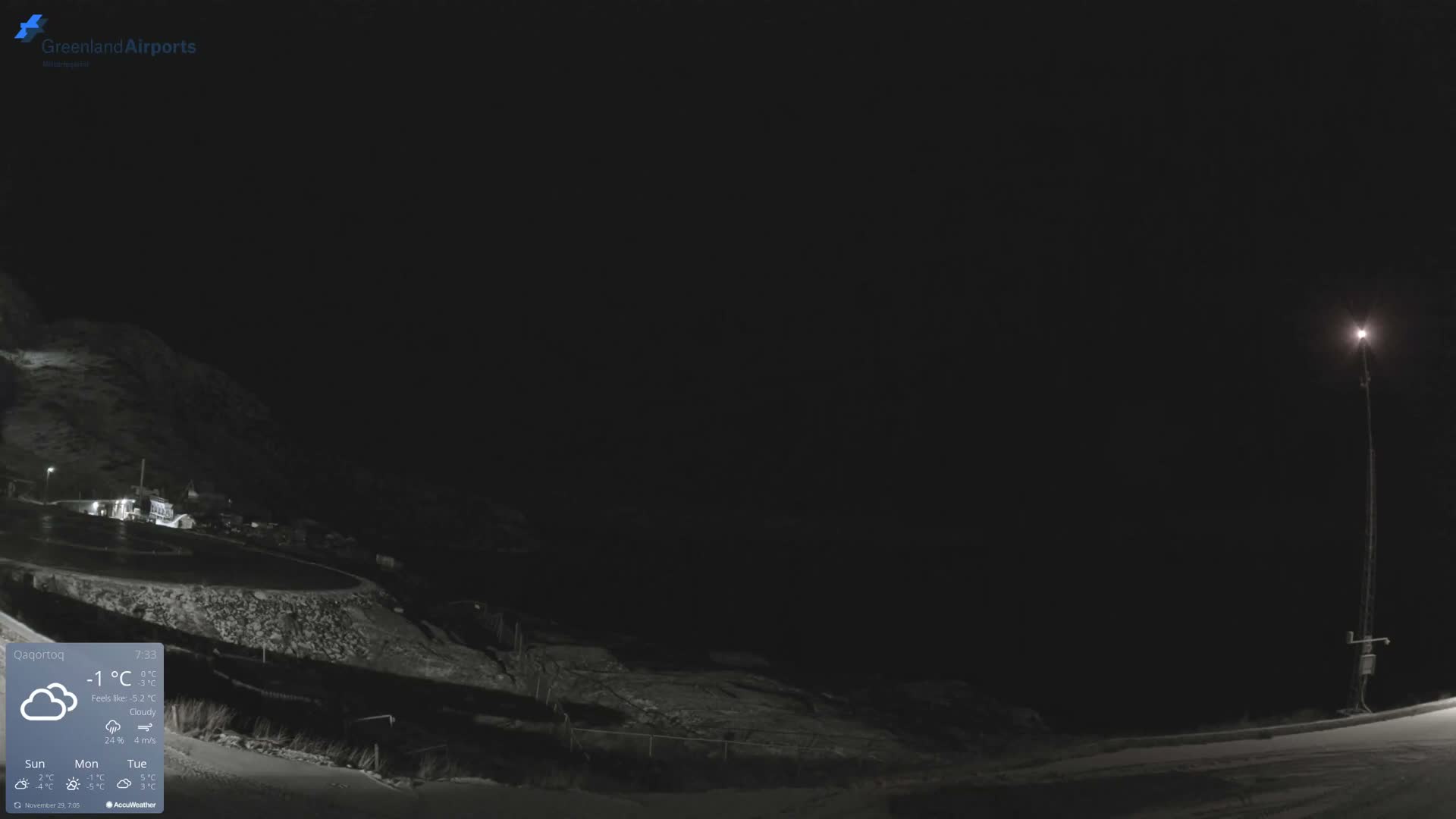 The image depicts a dark, snowy landscape at night, with distant illuminated buildings nestled against a mountain on the left and a prominent floodlight on a tall mast on the right, under a clear, cold sky.