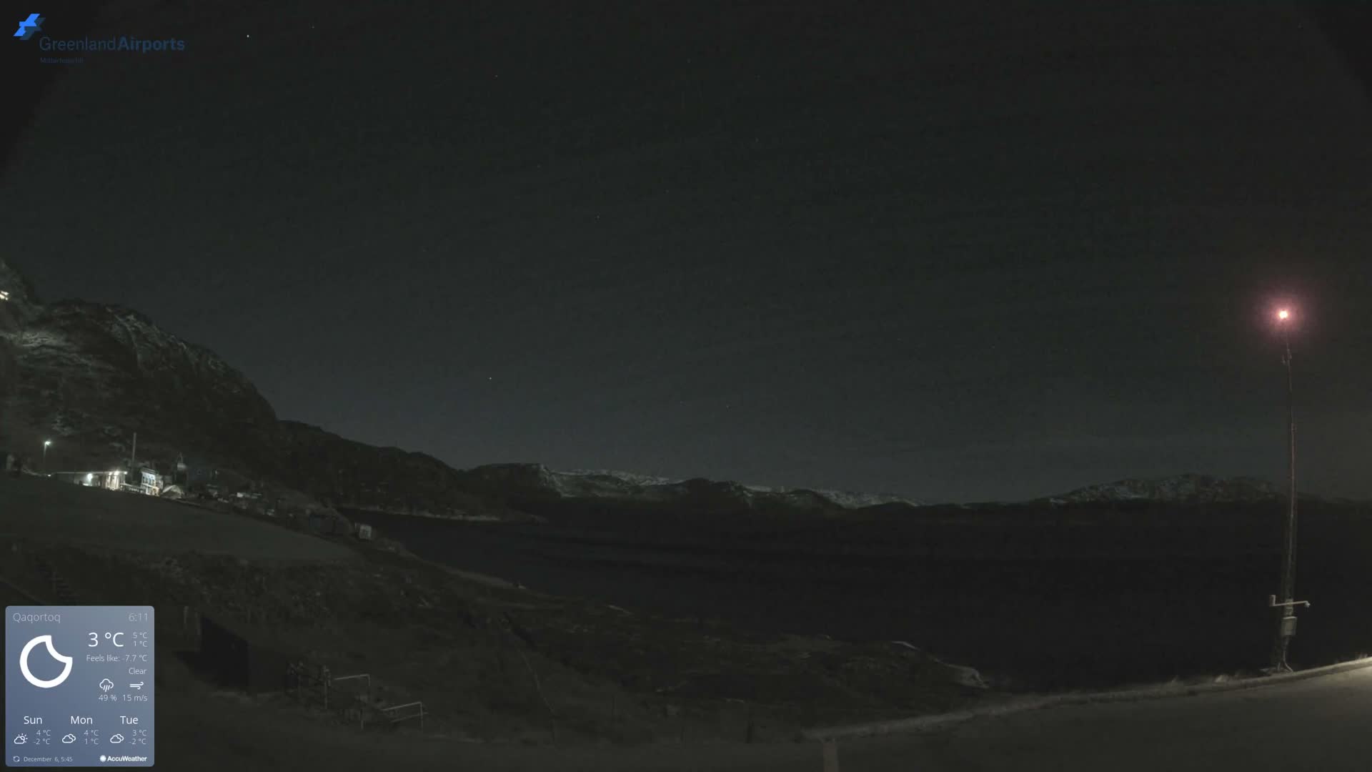 The image depicts a dark, clear night scene of a mountainous fjord landscape, with distant lights from buildings on a slope and a prominent red-glowing mast on the right.