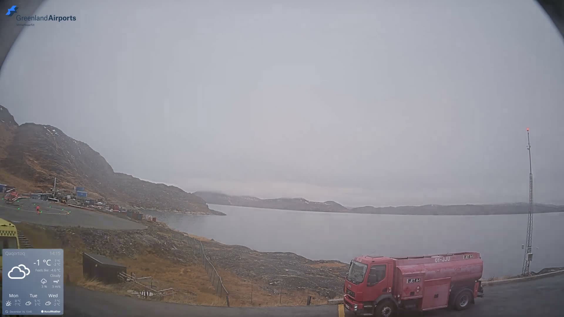 The image depicts a dark, clear night scene of a mountainous fjord landscape, with distant lights from buildings on a slope and a prominent red-glowing mast on the right.