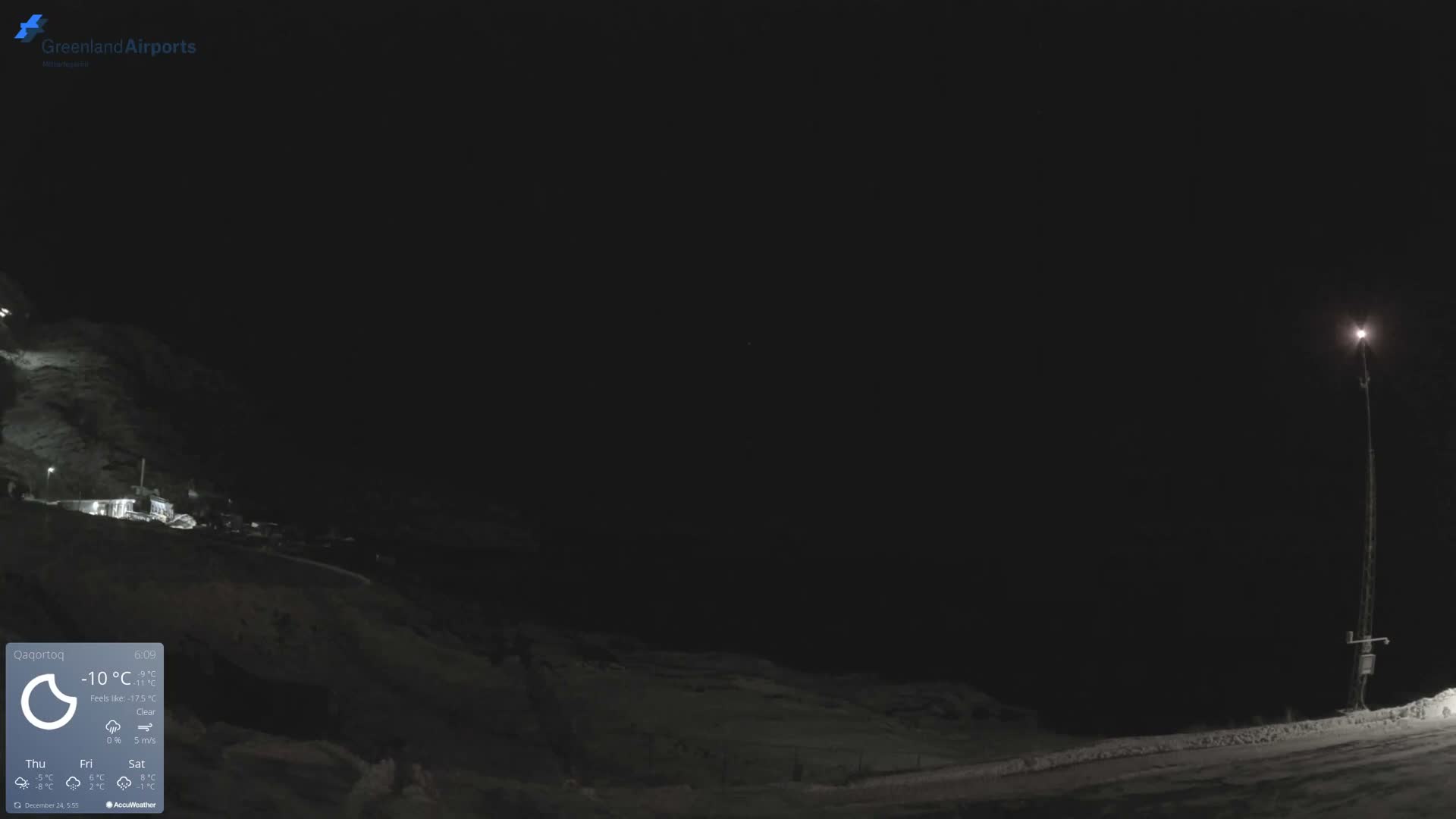A stark, dark night scene shows a tall illuminated light pole on the right and distant buildings on a snowy hillside to the left, under clear, cold conditions.