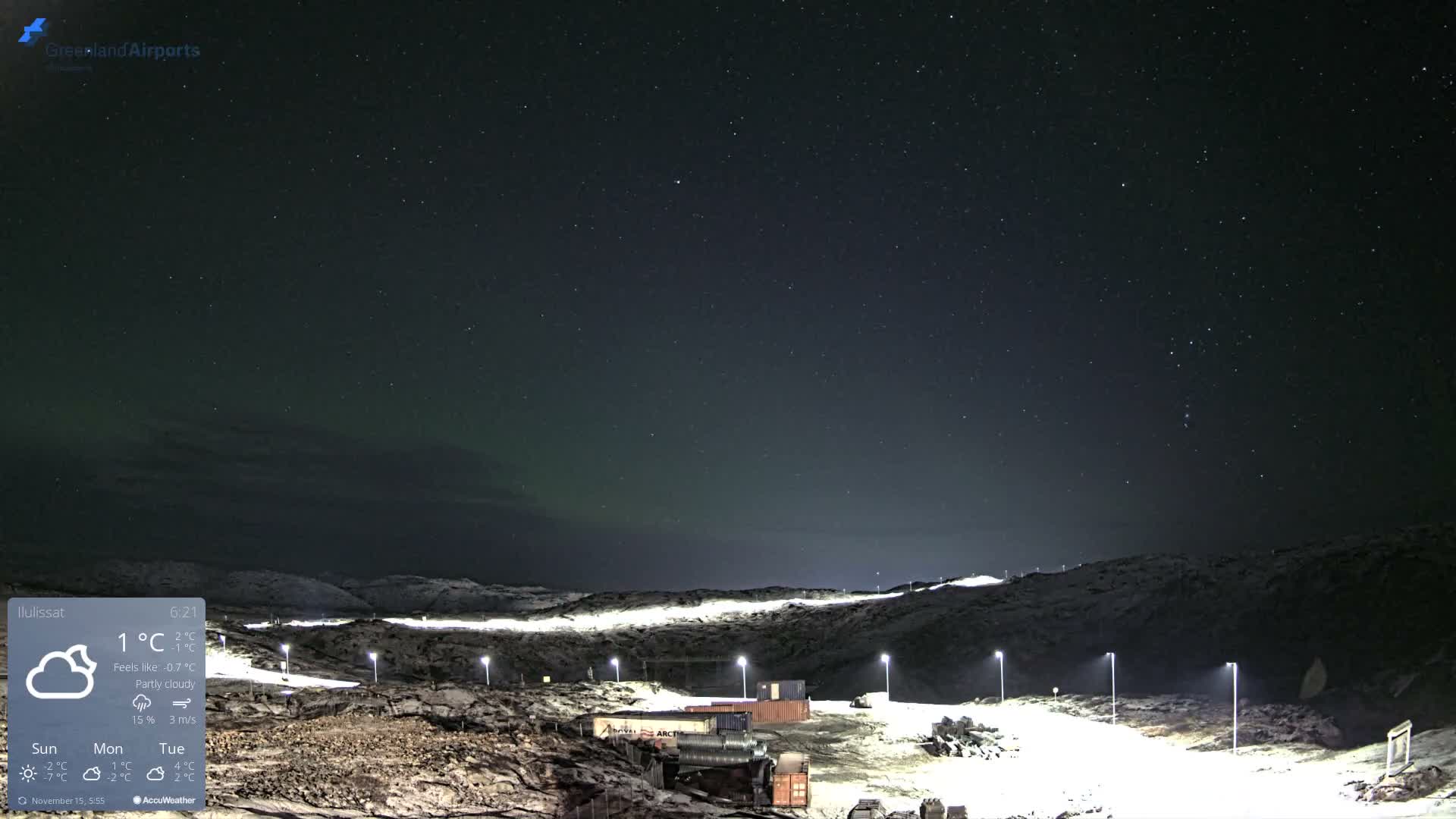 A cold, clear night reveals a snow-dusted, hilly landscape illuminated by streetlights and featuring scattered containers, all beneath a star-filled sky exhibiting a faint aurora borealis.