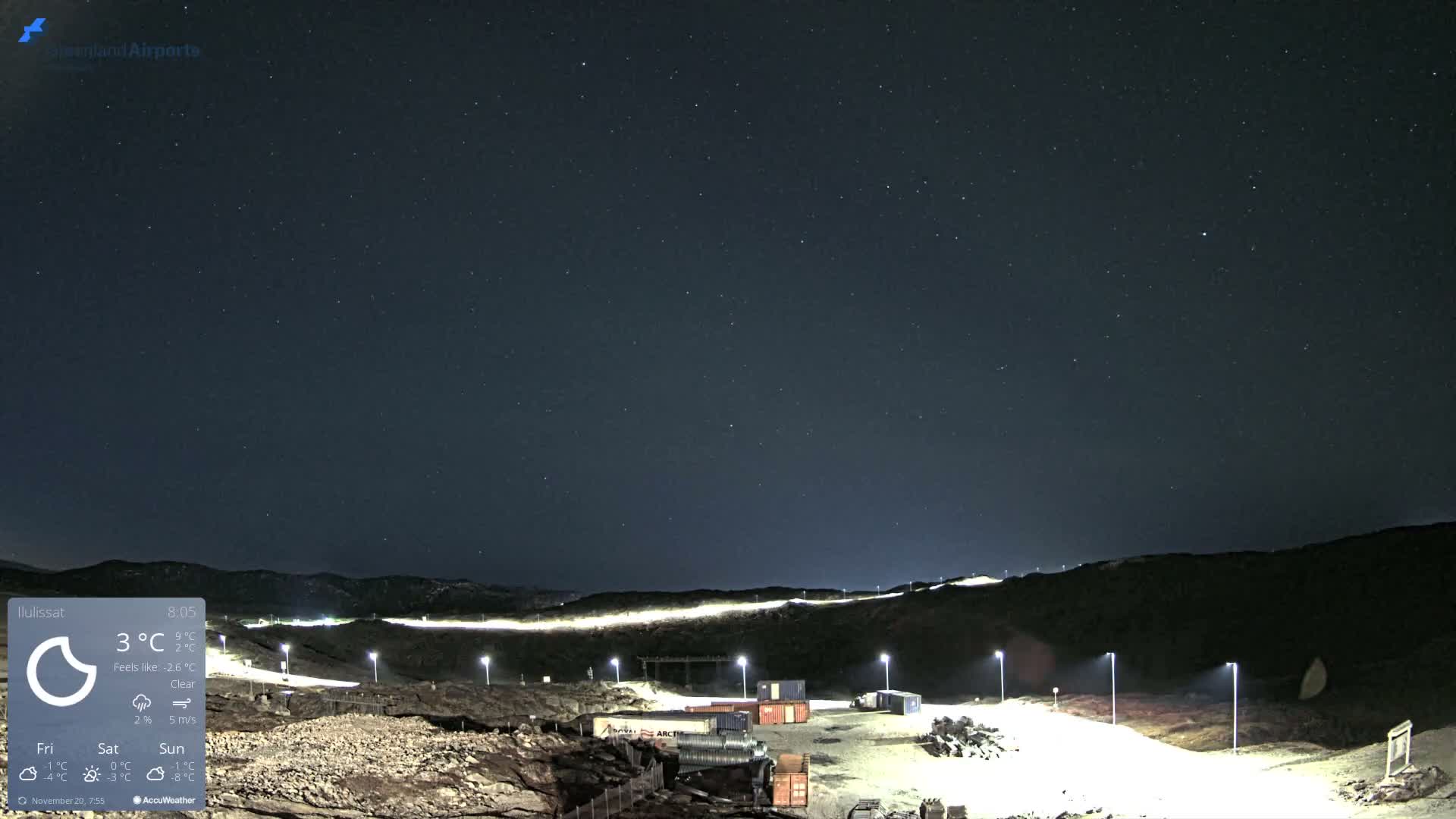 A clear, starry night sky hangs over a dark, hilly landscape, while the foreground reveals a brightly illuminated outdoor area with numerous streetlights, containers, and rocky terrain.