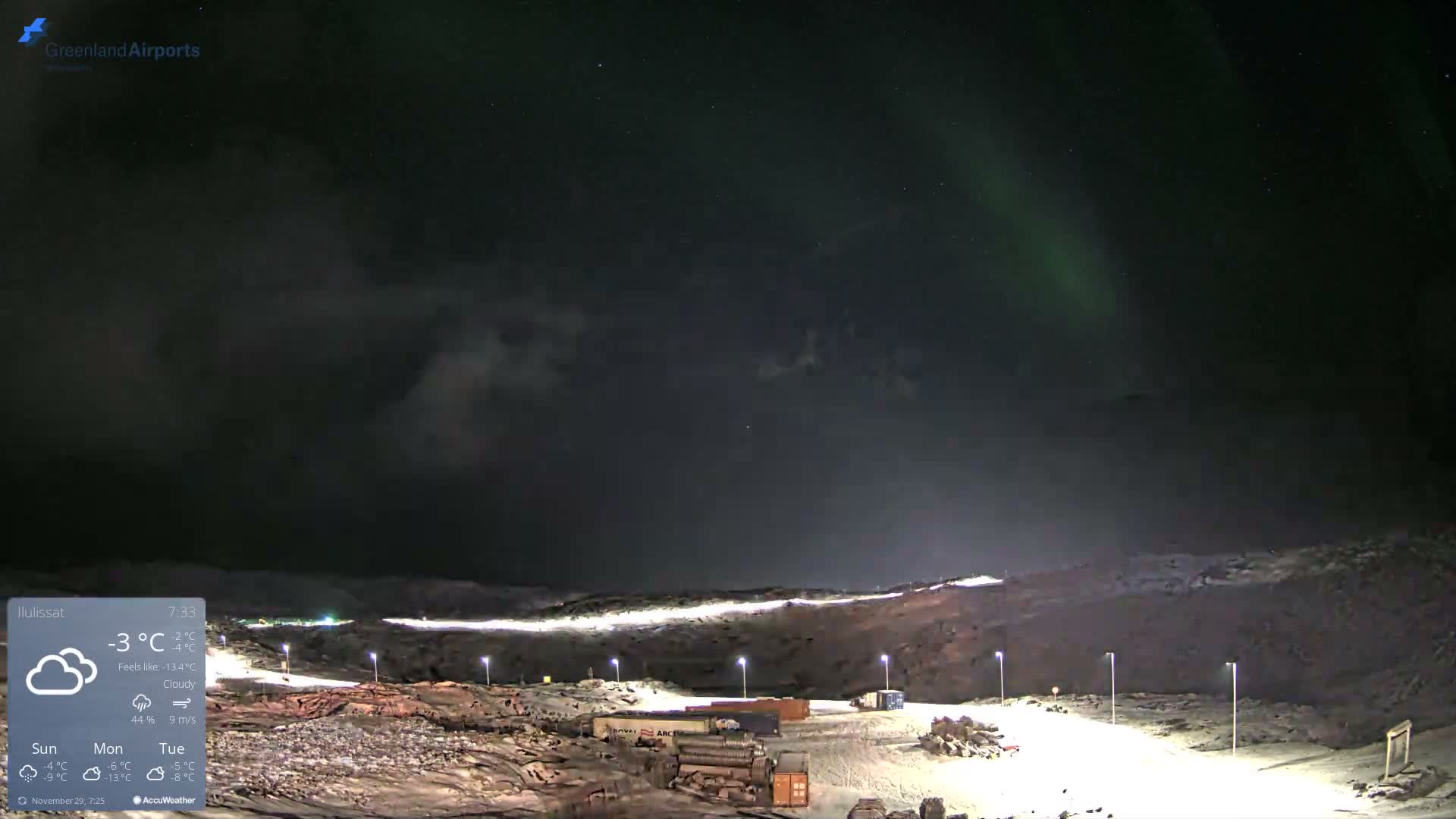 A vast, dark, and cloudy night sky with faint green aurora borealis hangs over a cold, snowy, and rugged landscape illuminated by numerous artificial lights.