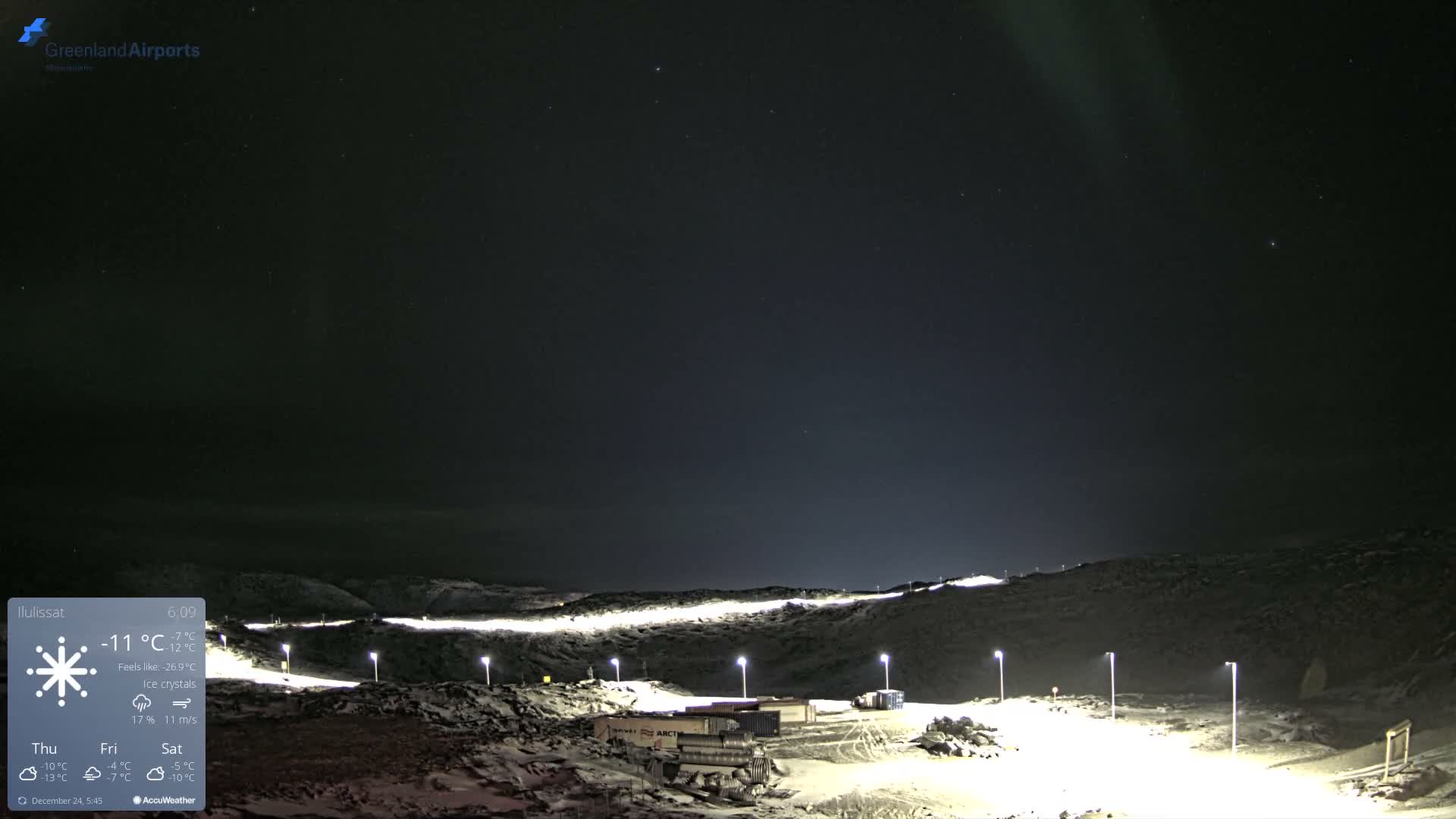 A clear, cold night reveals a snow-covered, hilly landscape illuminated by numerous streetlights, with structures alongside a winding road, all under a starry sky faintly touched by the aurora.