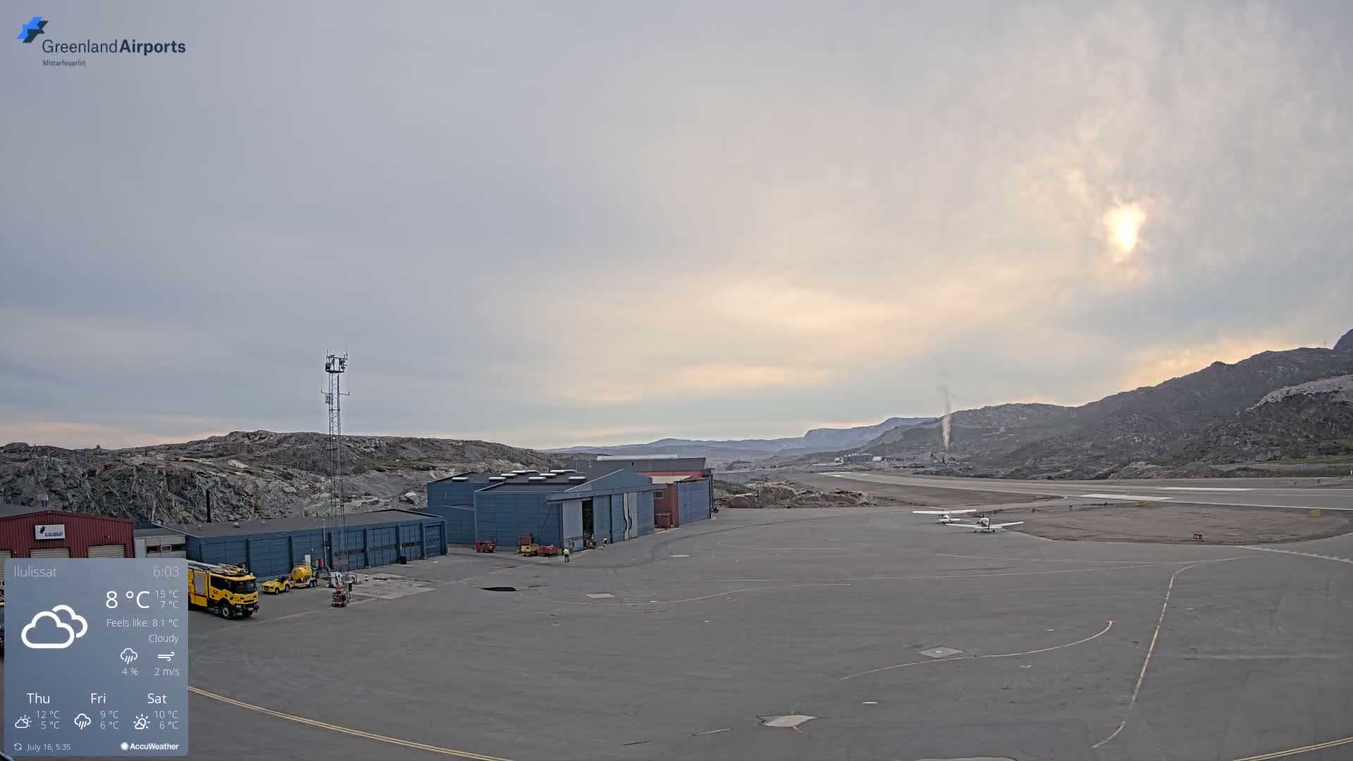 A small airport with several buildings and two small planes on the tarmac is situated under a cloudy but bright sky, near rocky hills.