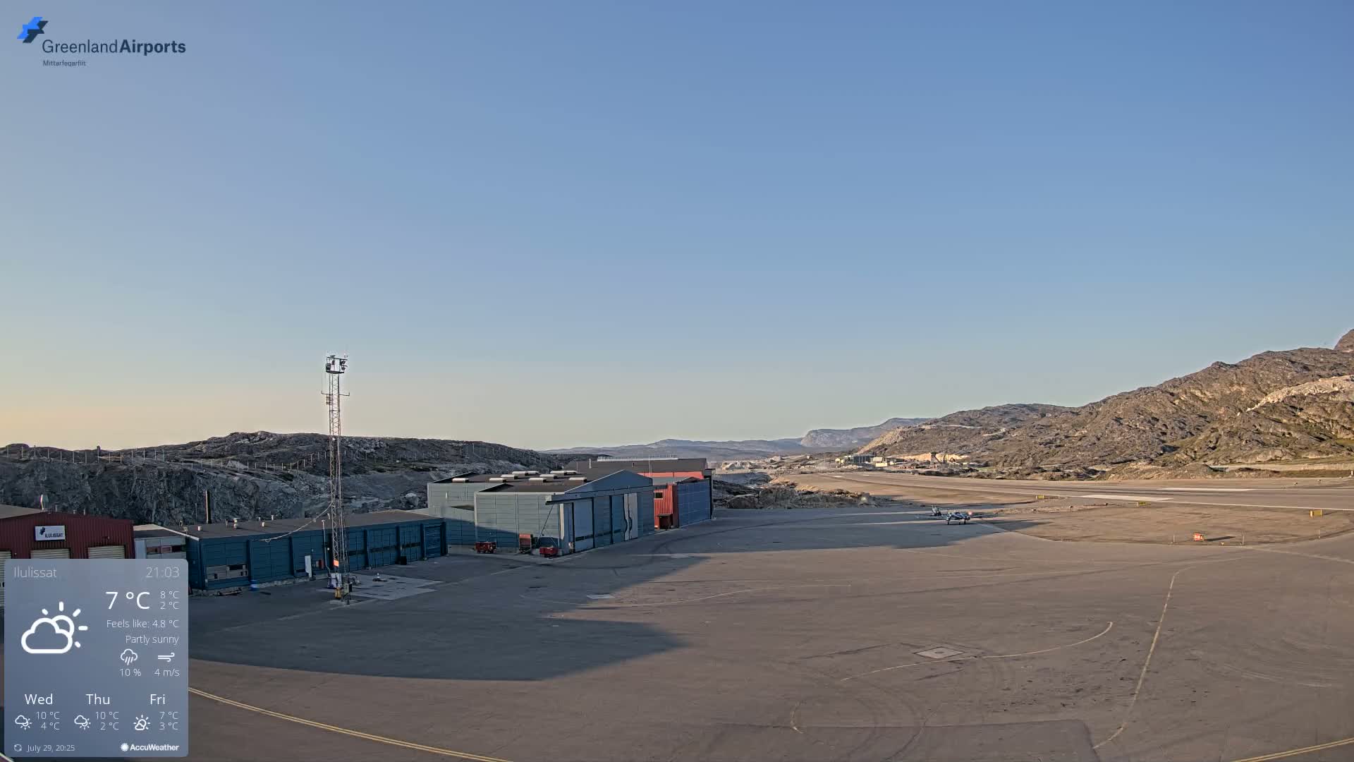 A small airport with several buildings and two small planes on the tarmac is situated under a cloudy but bright sky, near rocky hills.