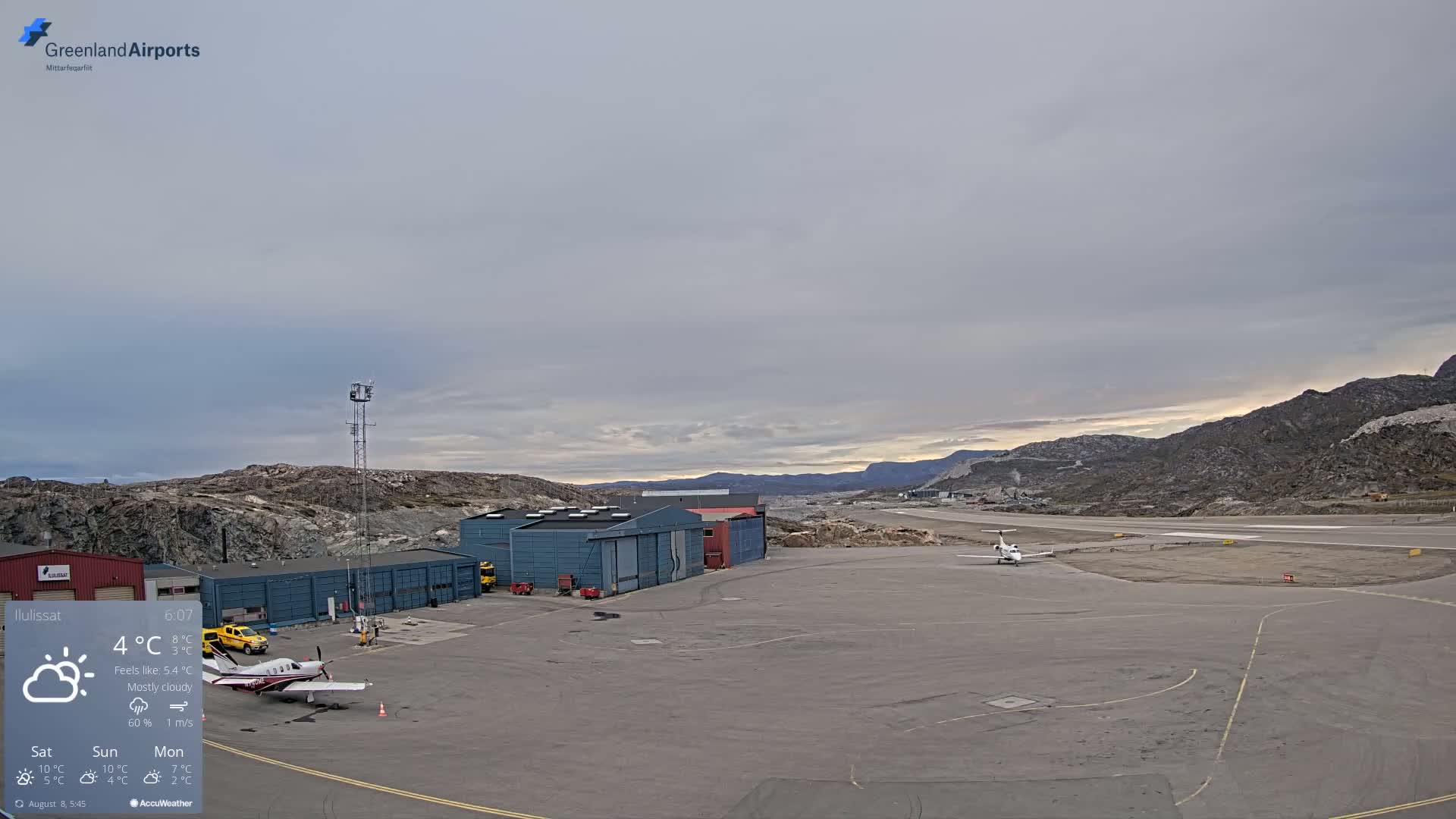 A mostly cloudy sky hangs over an airport tarmac with several small airplanes and airport buildings, nestled amongst rocky hills.