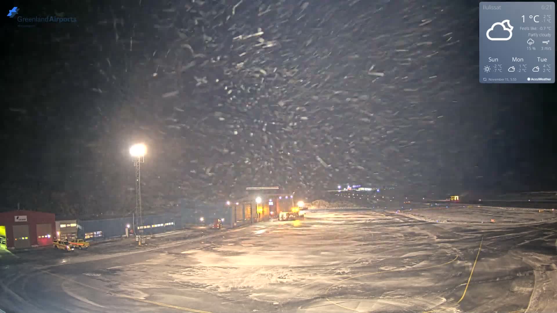 Heavy snow falls at night over a brightly lit, snow-covered airport tarmac, with maintenance buildings and vehicles visible in the foreground.