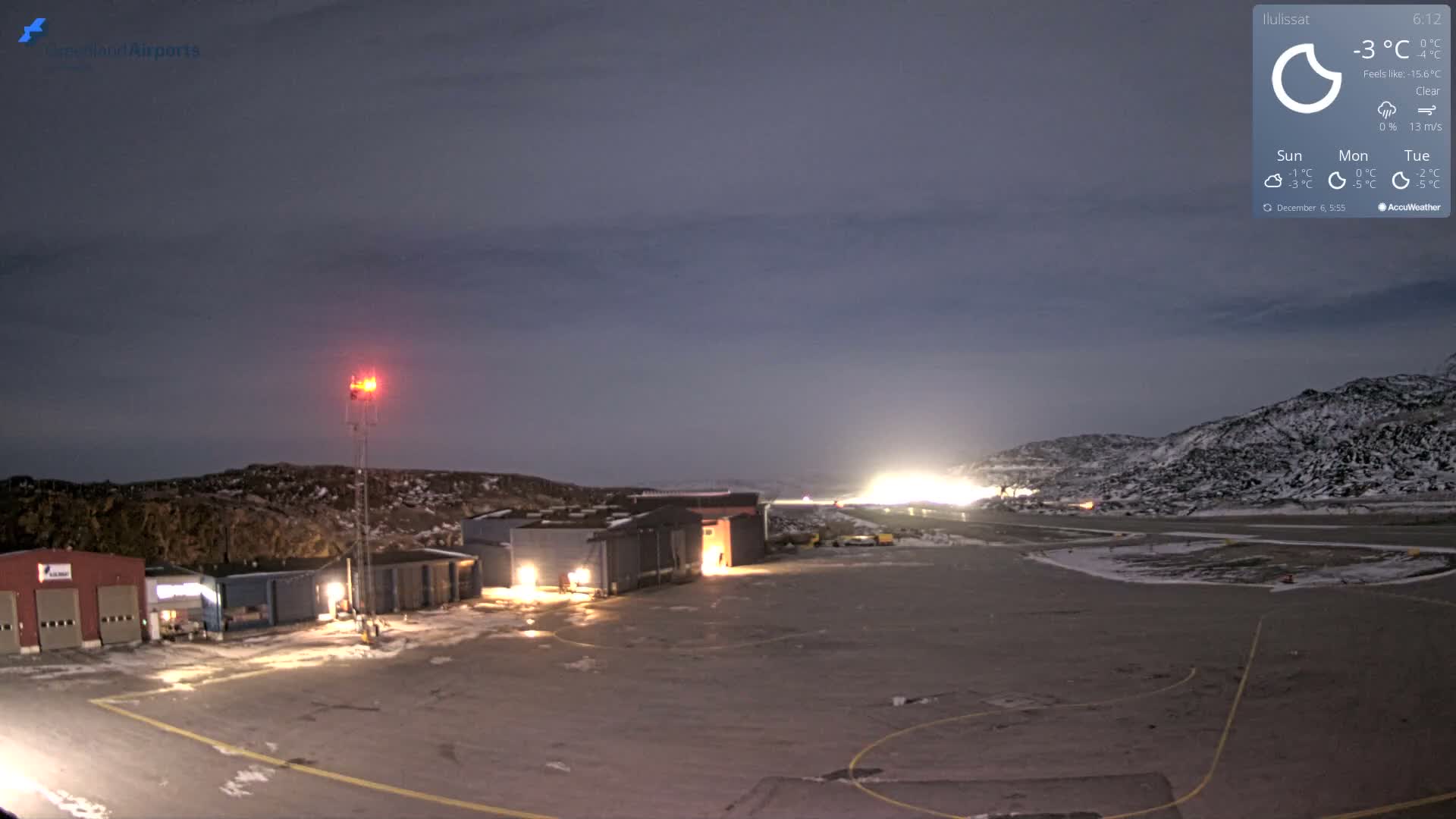 A clear, cold night view captures an airport scene with several low-rise buildings and a red beacon illuminated, a snow-dusted tarmac marked with yellow lines, and rugged, snowy hills under a dark sky, all dominated by a very bright light source on the distant horizon.
