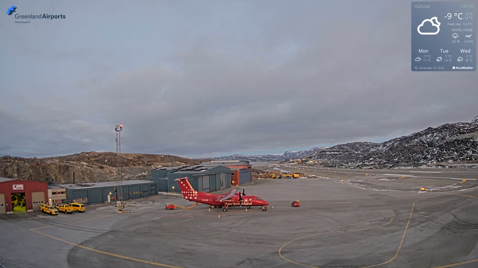 A clear, cold night view captures an airport scene with several low-rise buildings and a red beacon illuminated, a snow-dusted tarmac marked with yellow lines, and rugged, snowy hills under a dark sky, all dominated by a very bright light source on the distant horizon.