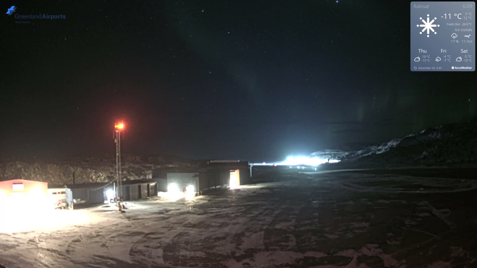 An expansive, snow-covered landscape at night reveals several brightly lit buildings and a communication tower with a red beacon, all beneath a clear, starry sky illuminated by the faint green glow of the aurora borealis, indicating freezing outdoor conditions.