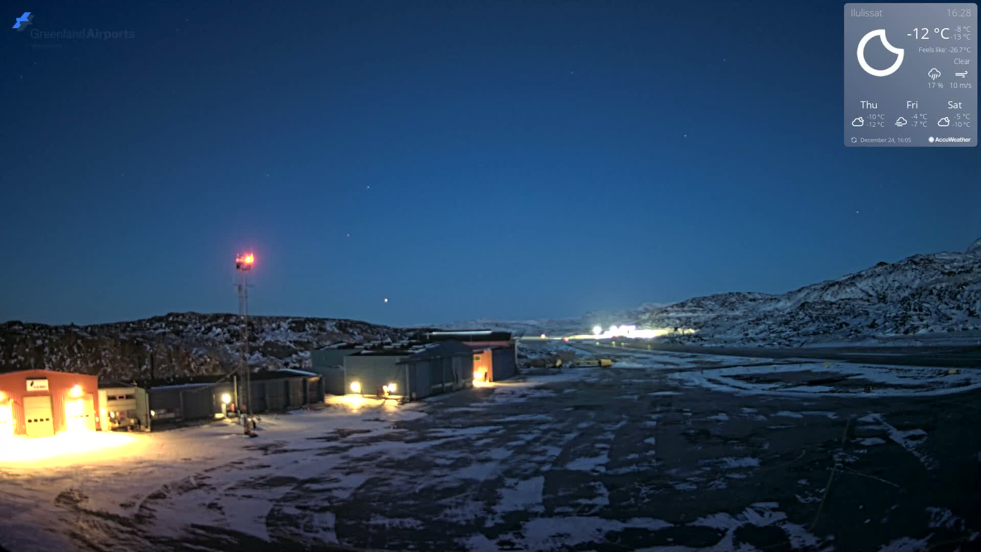 An expansive, snow-covered landscape at night reveals several brightly lit buildings and a communication tower with a red beacon, all beneath a clear, starry sky illuminated by the faint green glow of the aurora borealis, indicating freezing outdoor conditions.