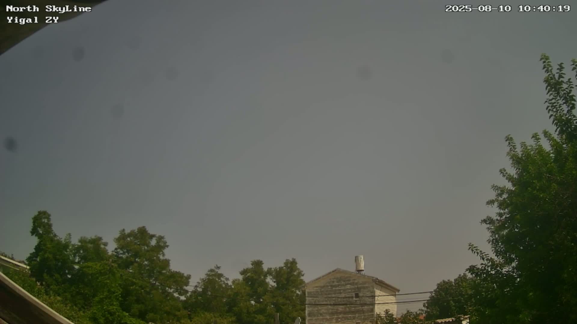 A hazy sky is visible above a small, light-colored building partially obscured by green trees.