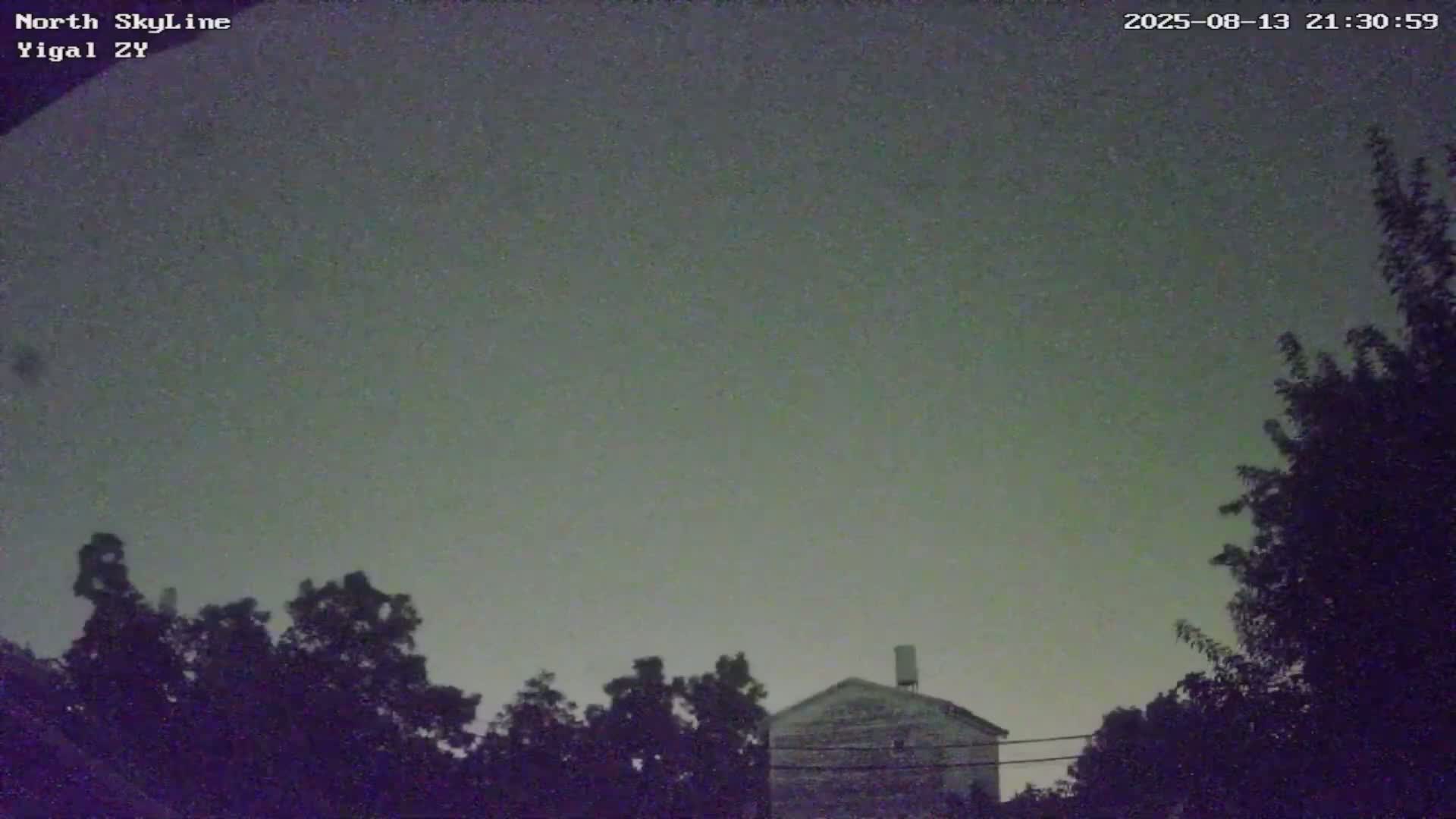 Under a dark, possibly overcast night sky, a small house is partially visible behind silhouetted trees.