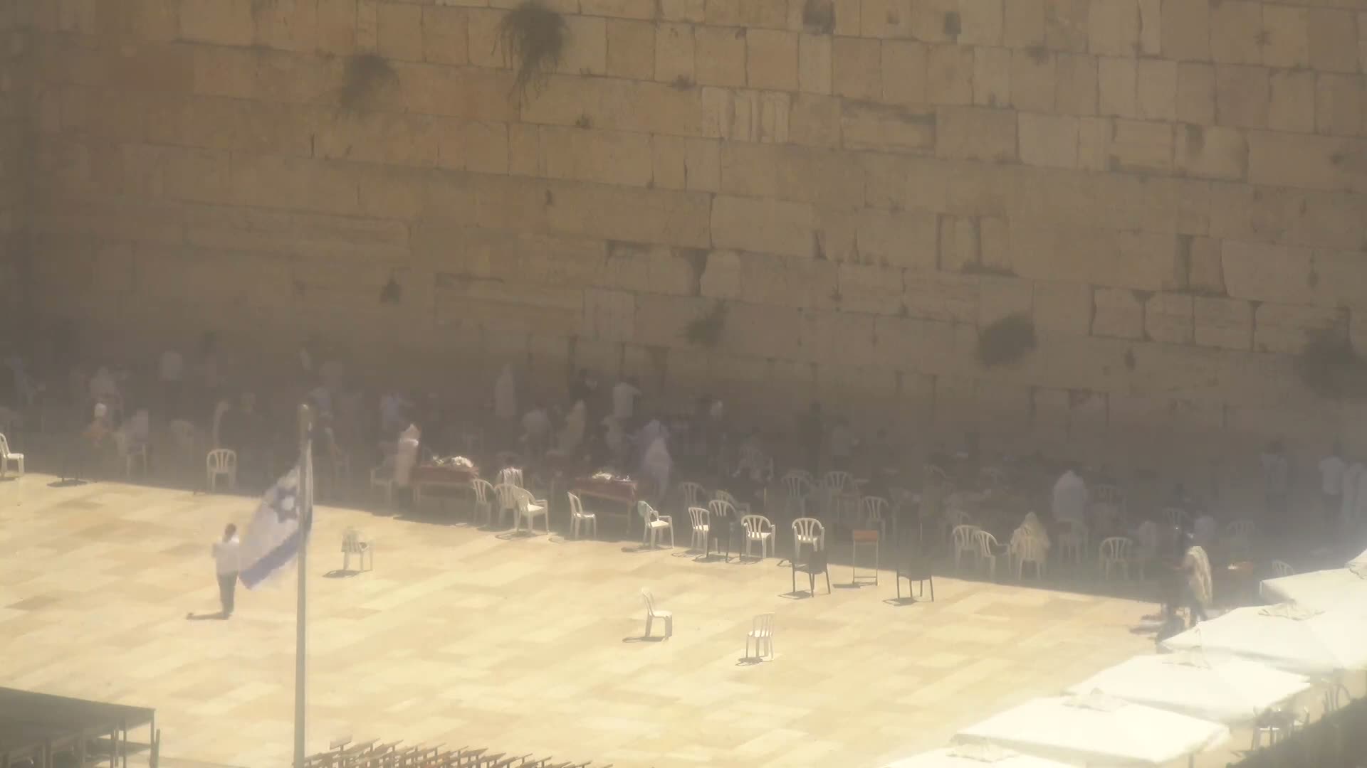 A large stone wall, under a hazy sun, has many people sitting in white plastic chairs in front of it.