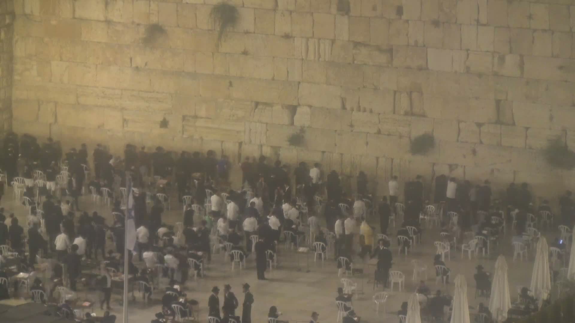 A large group of people in dark clothing are gathered in front of a tall, light-beige stone wall in foggy conditions.