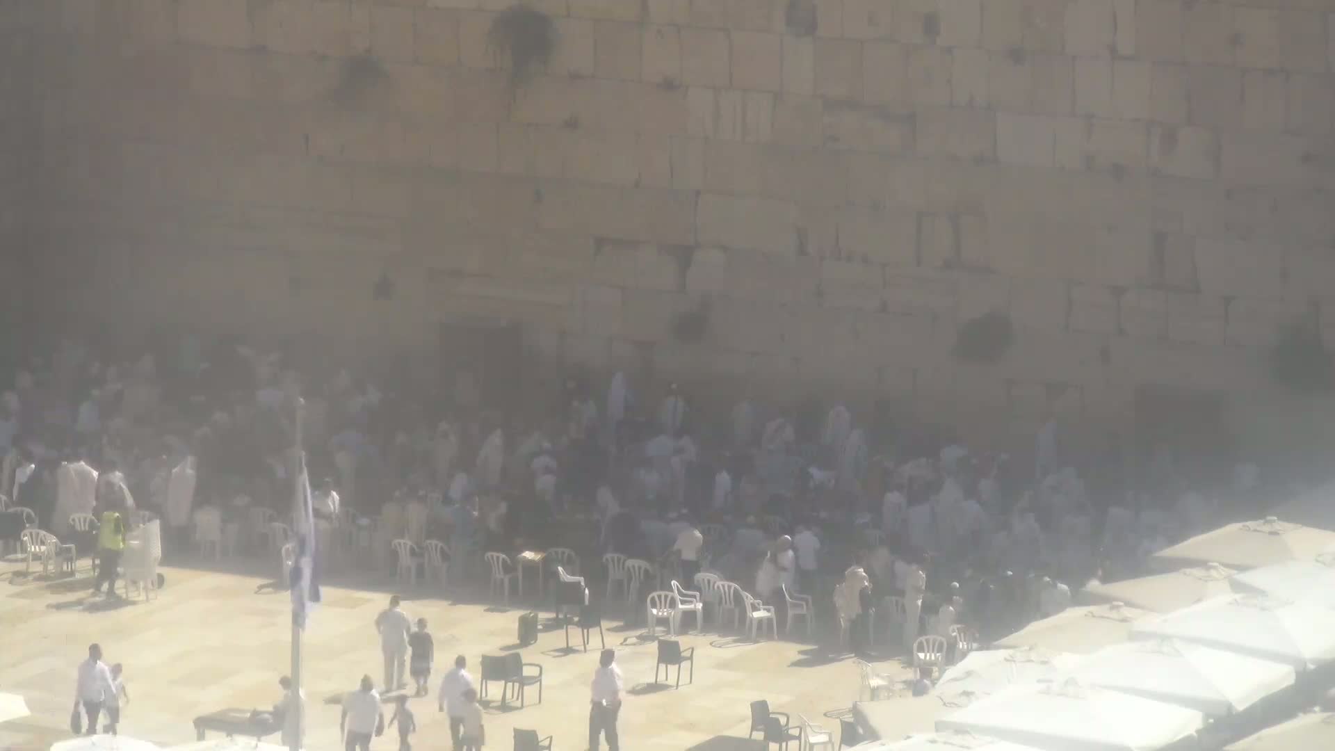 A hazy, sunny day shows many people gathered near a large stone wall, with some sitting in white plastic chairs and others standing.