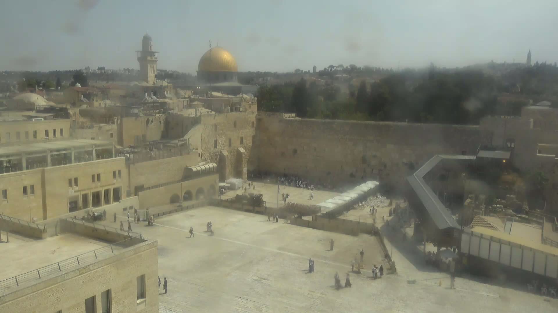 A hazy, high-angle view shows the Western Wall plaza in Jerusalem, with the Dome of the Rock visible in the background.