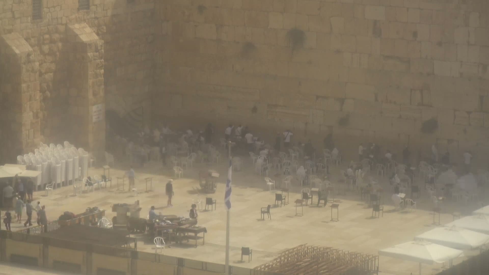 A hazy, outdoor view shows a large stone wall with many people sitting in white plastic chairs in front of it, some standing around tables and chairs in the plaza.