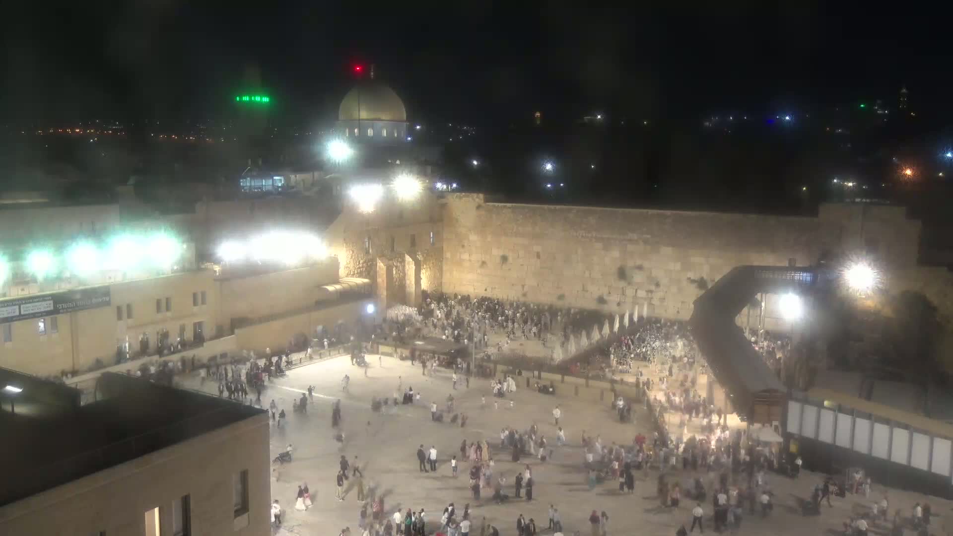 A nighttime, hazy view shows a large crowd gathered in a plaza in front of a high stone wall, with a gold-domed structure visible in the background.