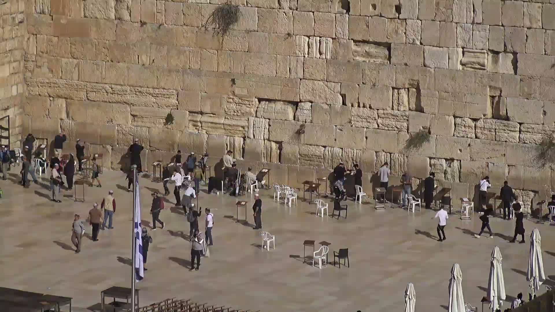 A nighttime, hazy view shows a large crowd gathered in a plaza in front of a high stone wall, with a gold-domed structure visible in the background.