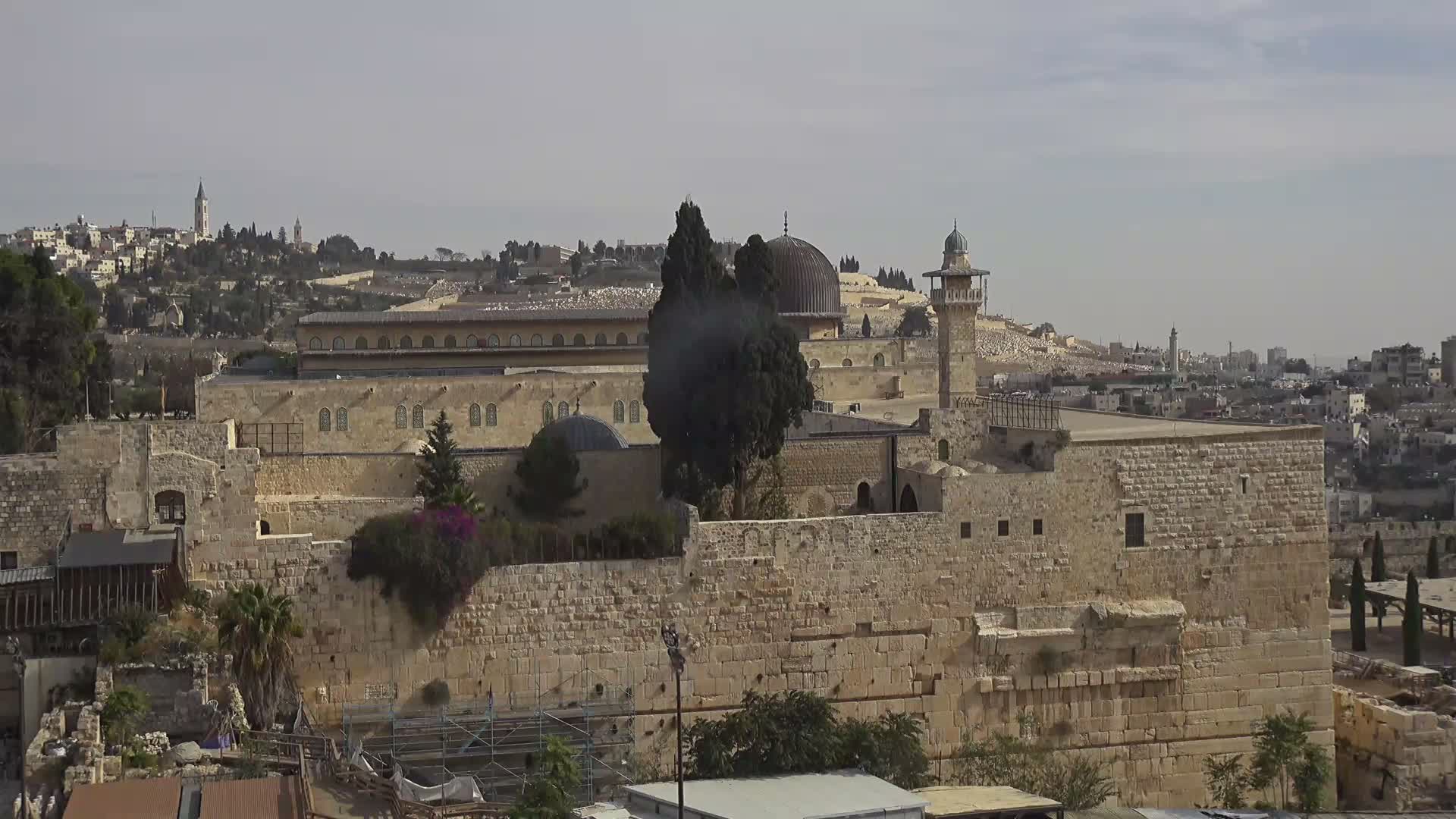 On a hazy day, an elevated panoramic view showcases the historic Al-Aqsa Mosque with its dark dome and minaret, surrounded by ancient stone walls and buildings, extending to a bustling cityscape and hills in the background.