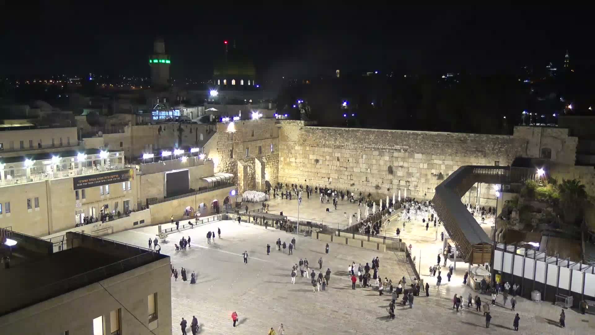 The image captures a panoramic view of an ancient city's stone architecture, featuring prominent domes and a minaret, with cityscapes and hills extending into the background under a bright, partly cloudy sky.