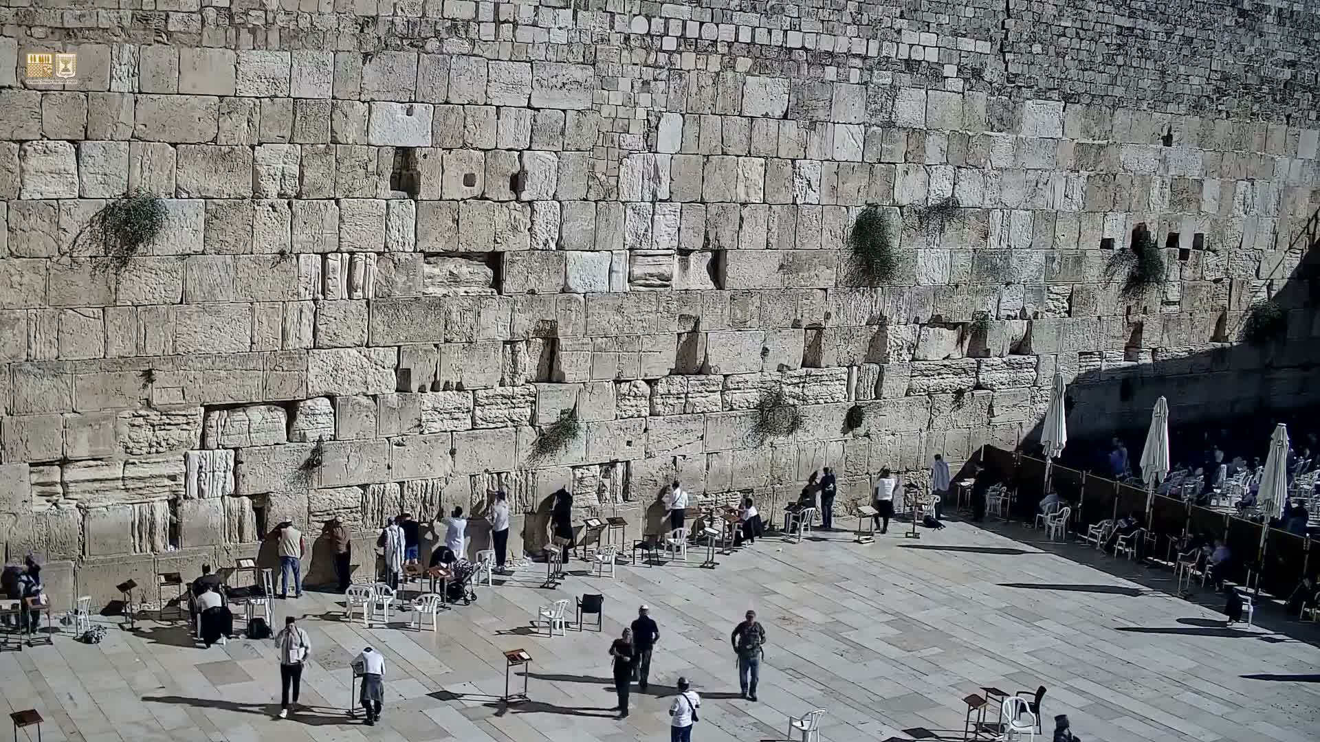 A large crowd gathers on a bright, sunny day at the ancient stone Western Wall, with many people standing near the facade and others seated under white umbrellas in the sunlit plaza.
