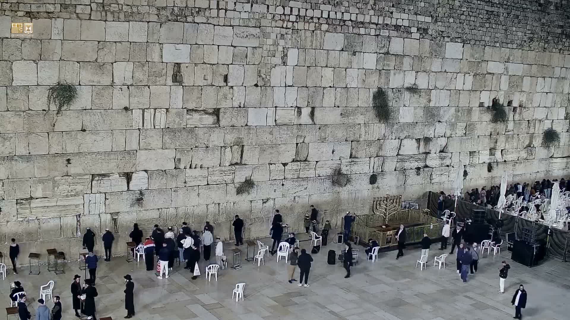 A large crowd gathers on a bright, sunny day at the ancient stone Western Wall, with many people standing near the facade and others seated under white umbrellas in the sunlit plaza.