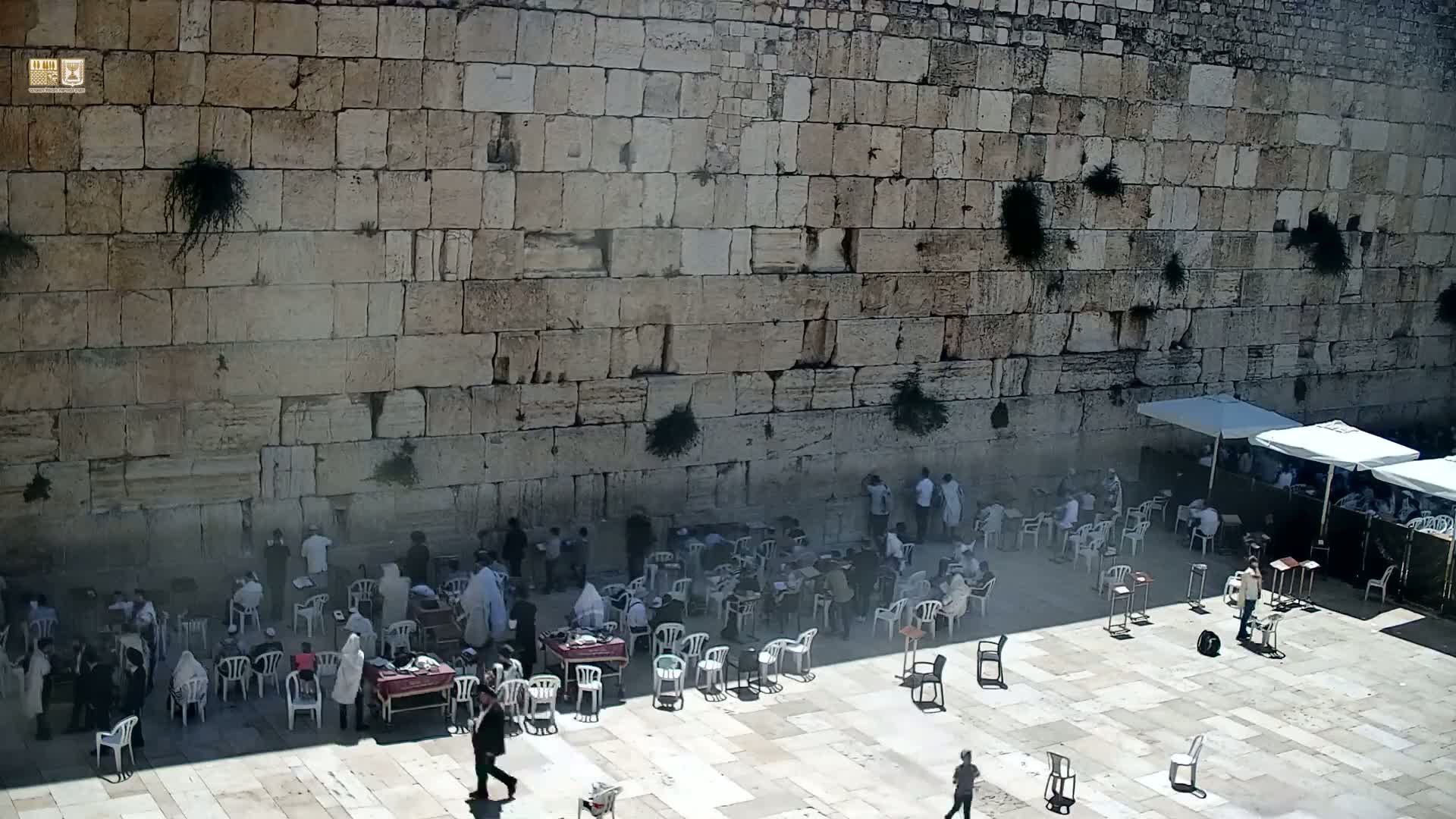A large stone wall is surrounded by people sitting in white plastic chairs under a sunny sky.