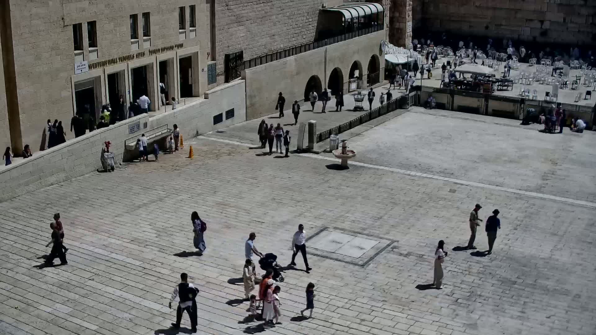 A large stone plaza, under sunny conditions, is populated with numerous people walking and sitting, with some gathered near a building and a water fountain.