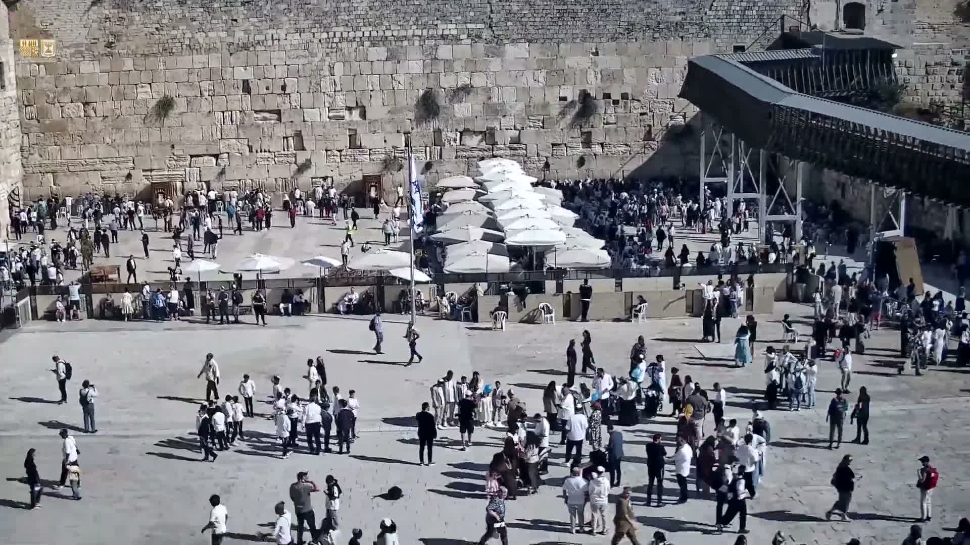 The outdoor plaza of the Western Wall is bustling with numerous people on a clear, sunny day, featuring many white umbrellas and an elevated walkway on the right.