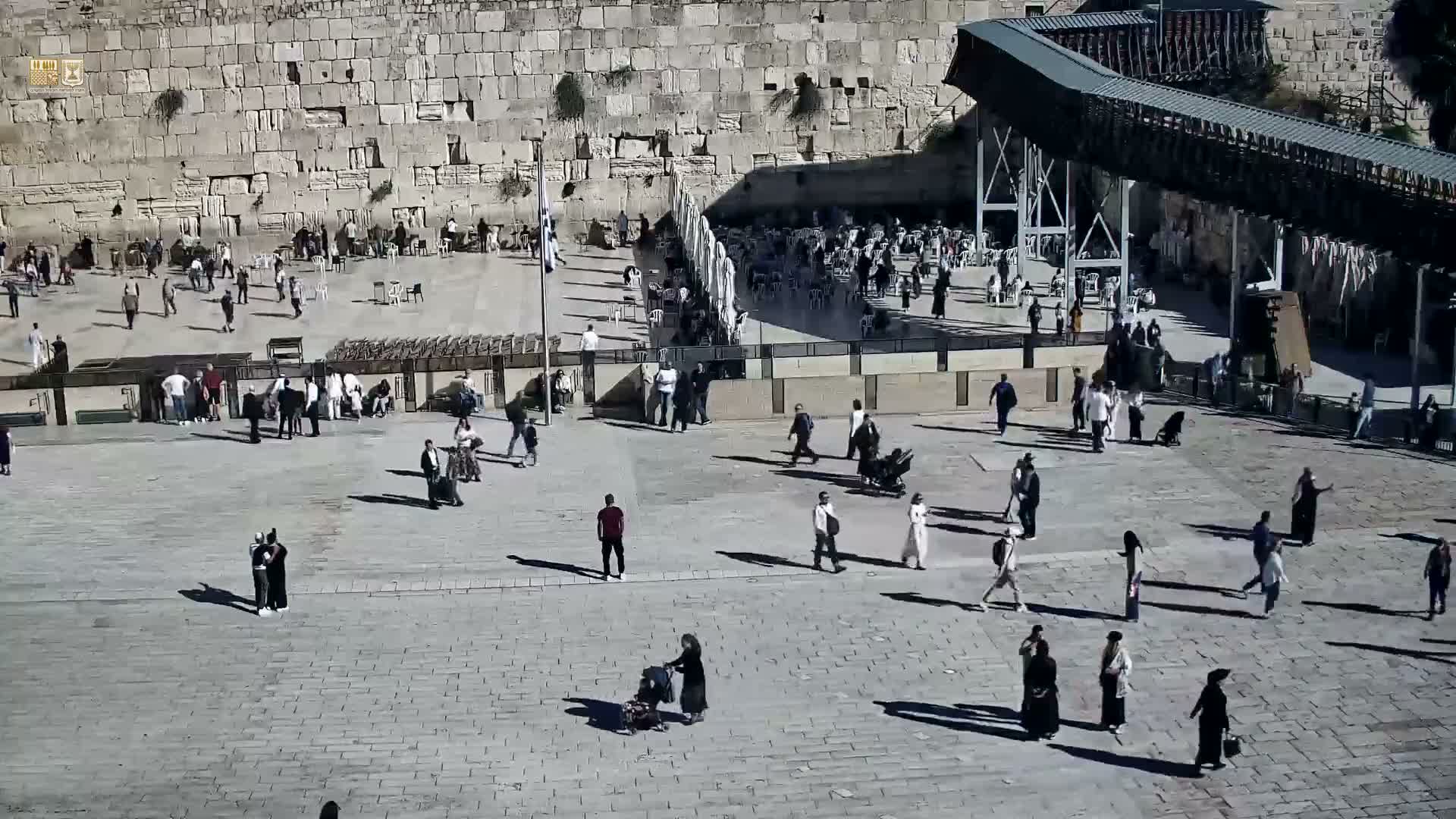 The outdoor plaza of the Western Wall is bustling with numerous people on a clear, sunny day, featuring many white umbrellas and an elevated walkway on the right.