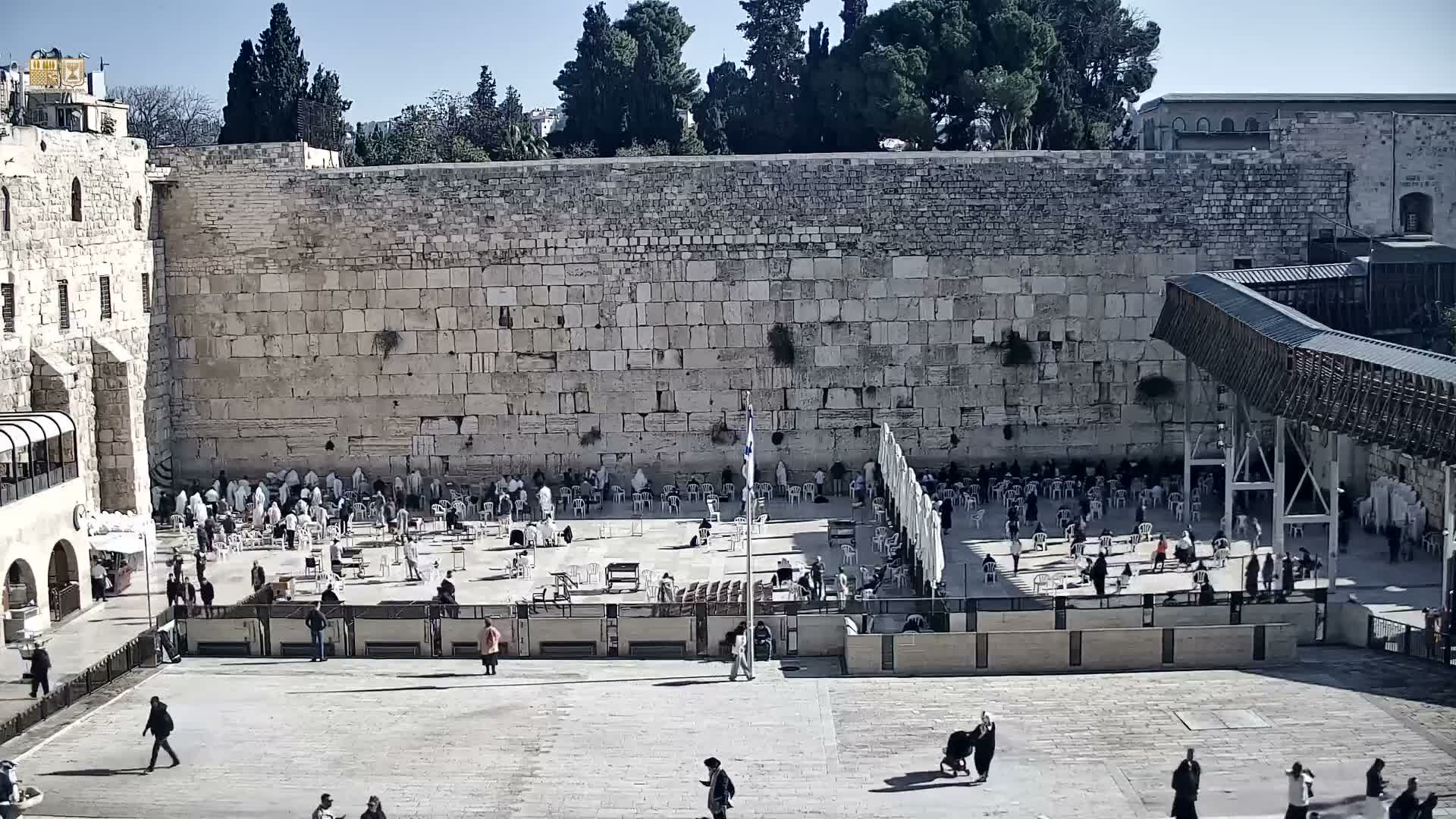 On a bright, clear day, numerous people are gathered and praying in the paved plaza before the ancient Western Wall, with a flagpole, temporary seating, and a modern walkway visible under a clear blue sky.
